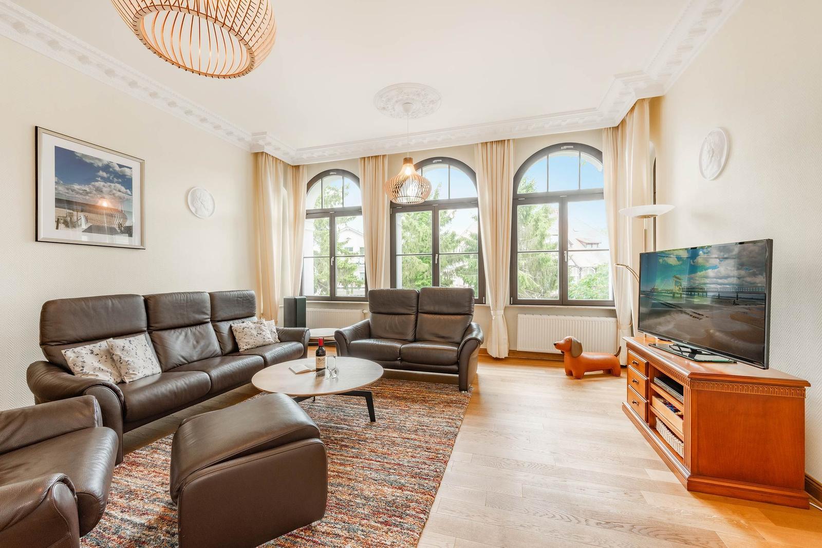 Wohnzimmer mit Ledersofa, Fernseher und großen Fenstern.