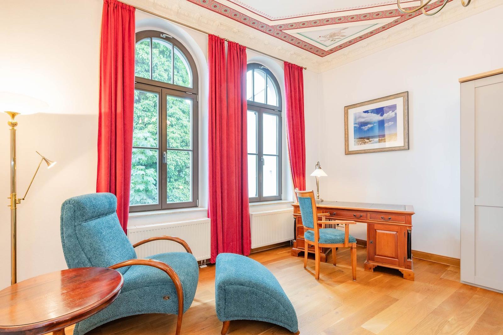 Ein Raum mit blauem Sessel, Schreibtisch und roten Vorhängen an den Fenstern.