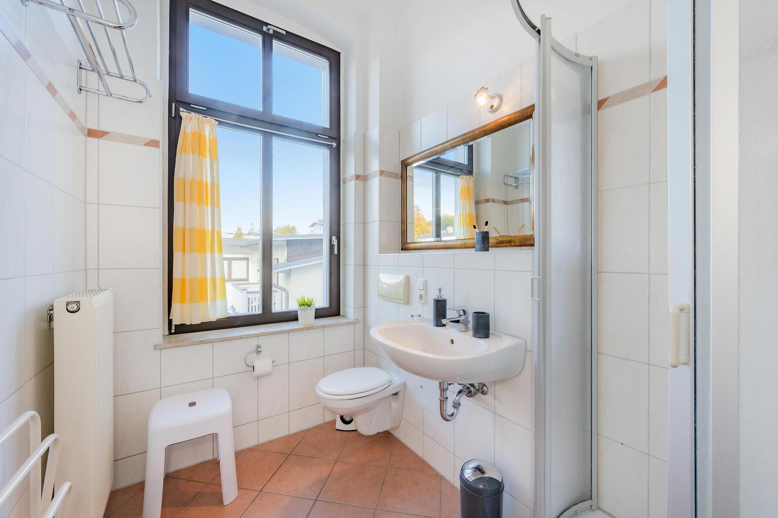 Badezimmer mit Dusche, Waschbecken, Toilette und Fenster mit gelb-weißem Vorhang.