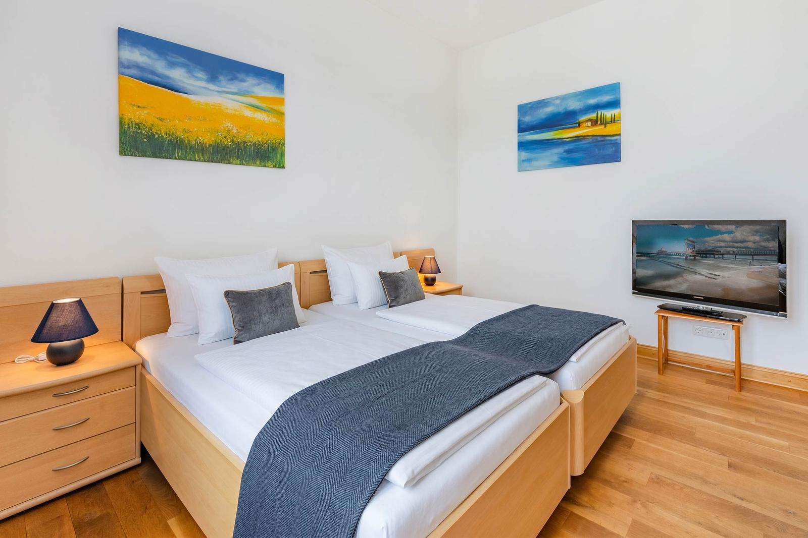 Zimmer mit Doppelbett, Nachttisch, Fernseher und Wandbildern.