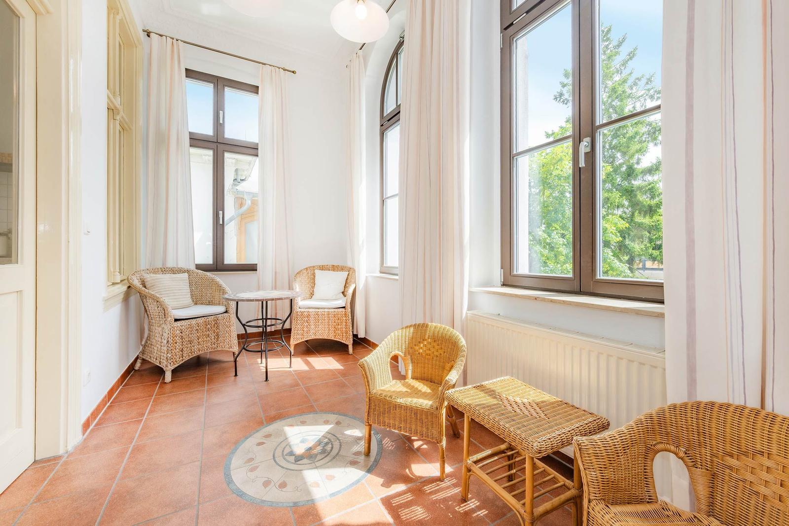 Raum mit Rattanmöbeln, Terrakottaboden und großen Fenstern mit Blick auf Bäume.