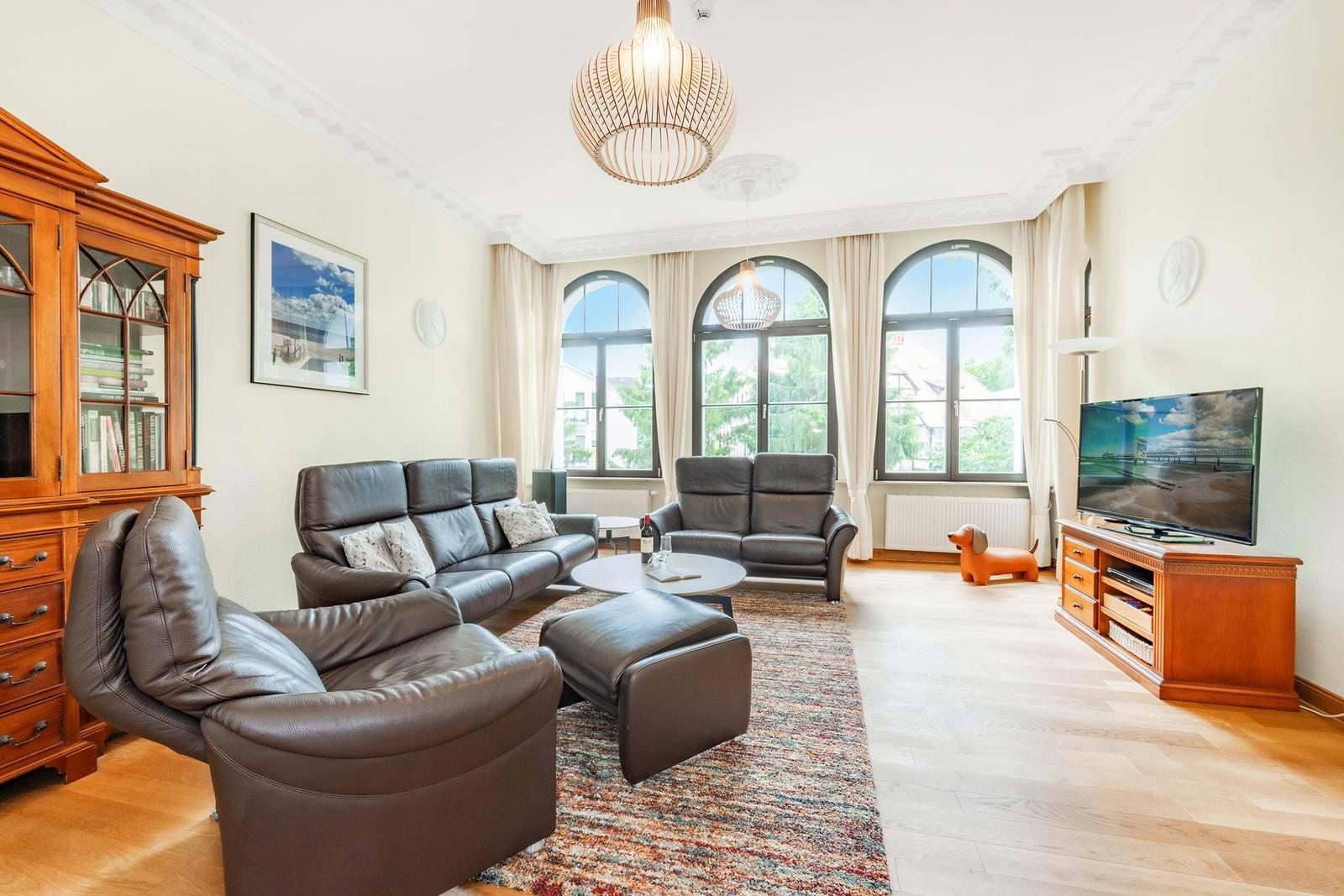 Wohnzimmer mit Ledersofa, Fernseher und drei Bogenfenstern mit Blick nach draußen.
