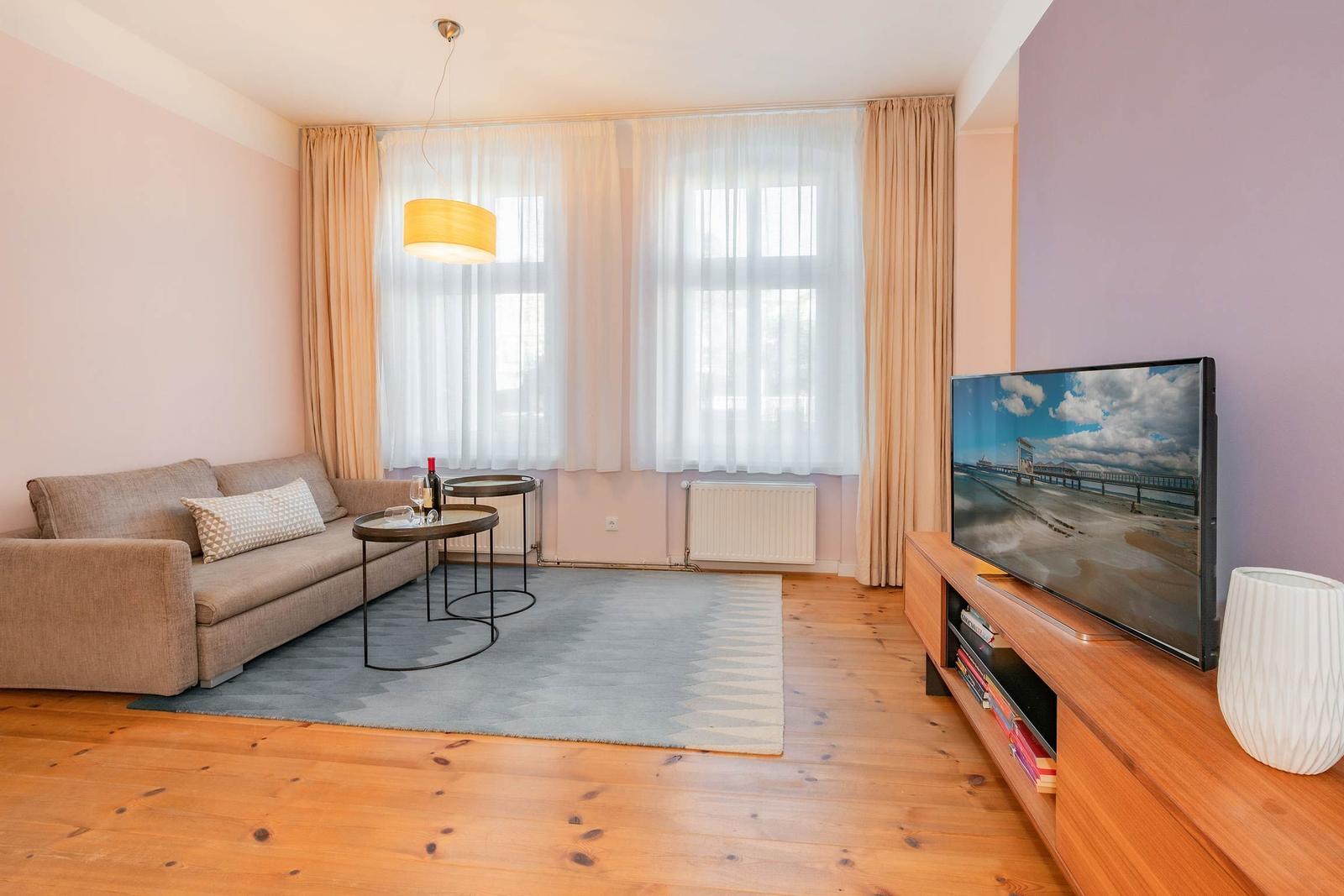 Wohnzimmer mit Sofa, TV und Holzboden. Fenster mit Vorhängen und Teppich.