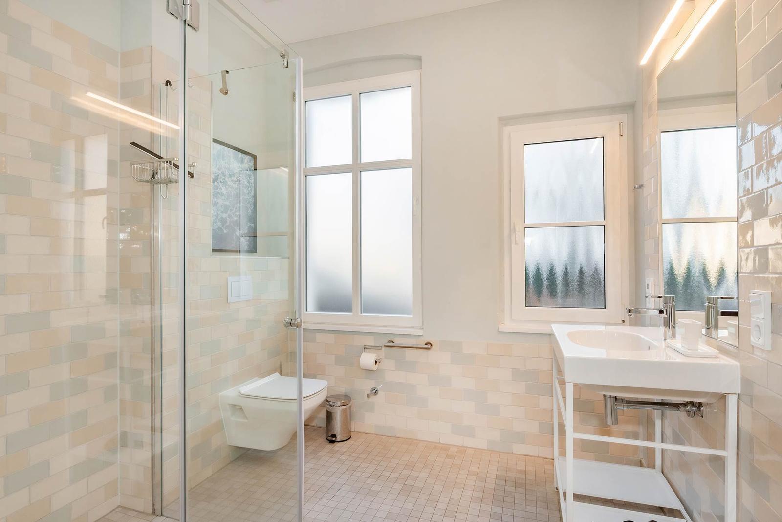 Badezimmer mit Dusche, Toilette und Waschbecken. Fenster mit getöntem Glas.