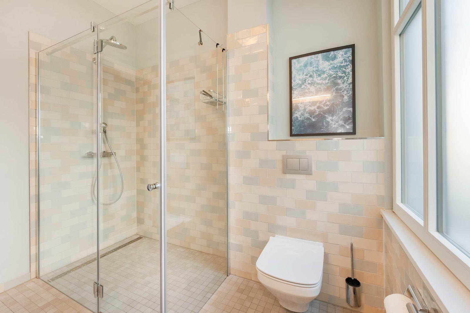Dusche mit Glaswand, Toilette und Fenster. Wand mit Fliesen und Bild.
