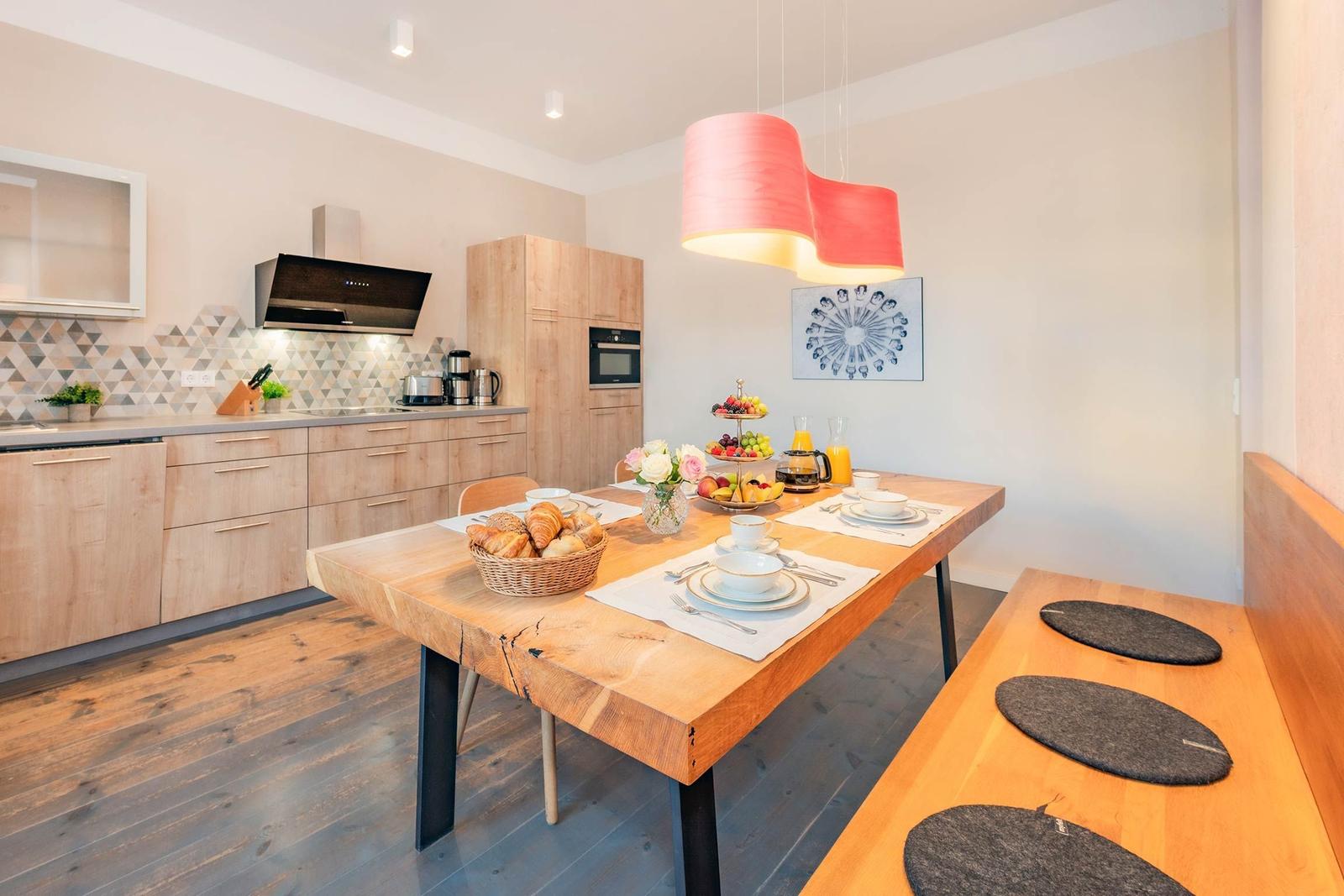 Essbereich mit Holztisch, Bank und Frühstückszubehör. Küche mit Holzschränken und Deckenleuchte.