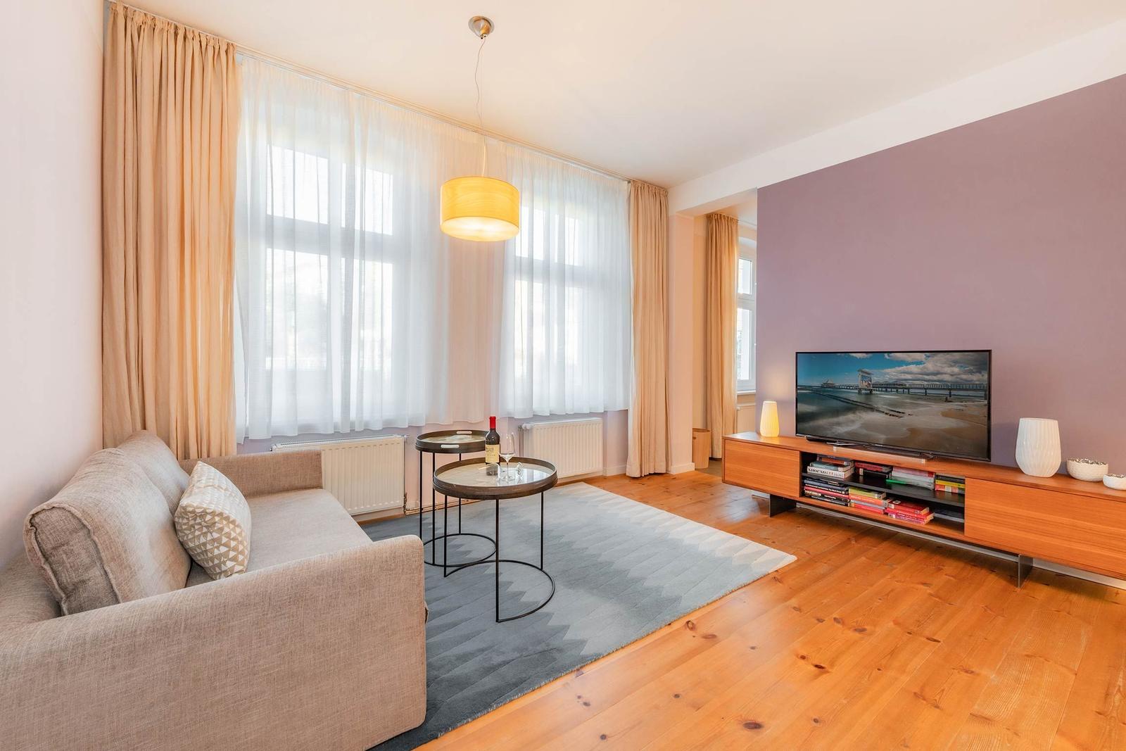 Wohnzimmer mit Sofa, TV, Tisch und großen Fenstern mit Vorhängen.