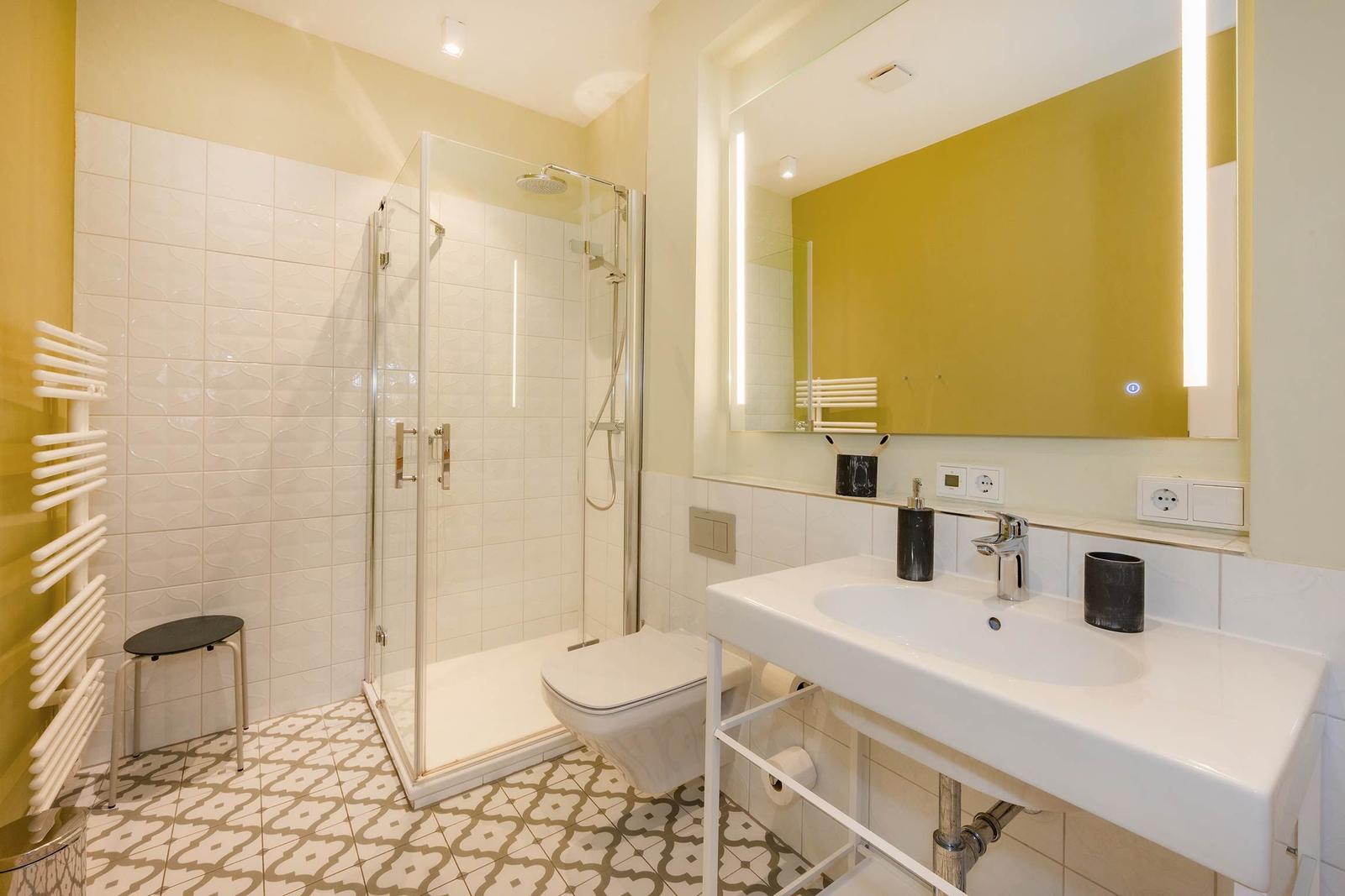 Badezimmer mit Dusche, Waschbecken und Toilette. Gelbe Wände und gemusterte Fliesen.
