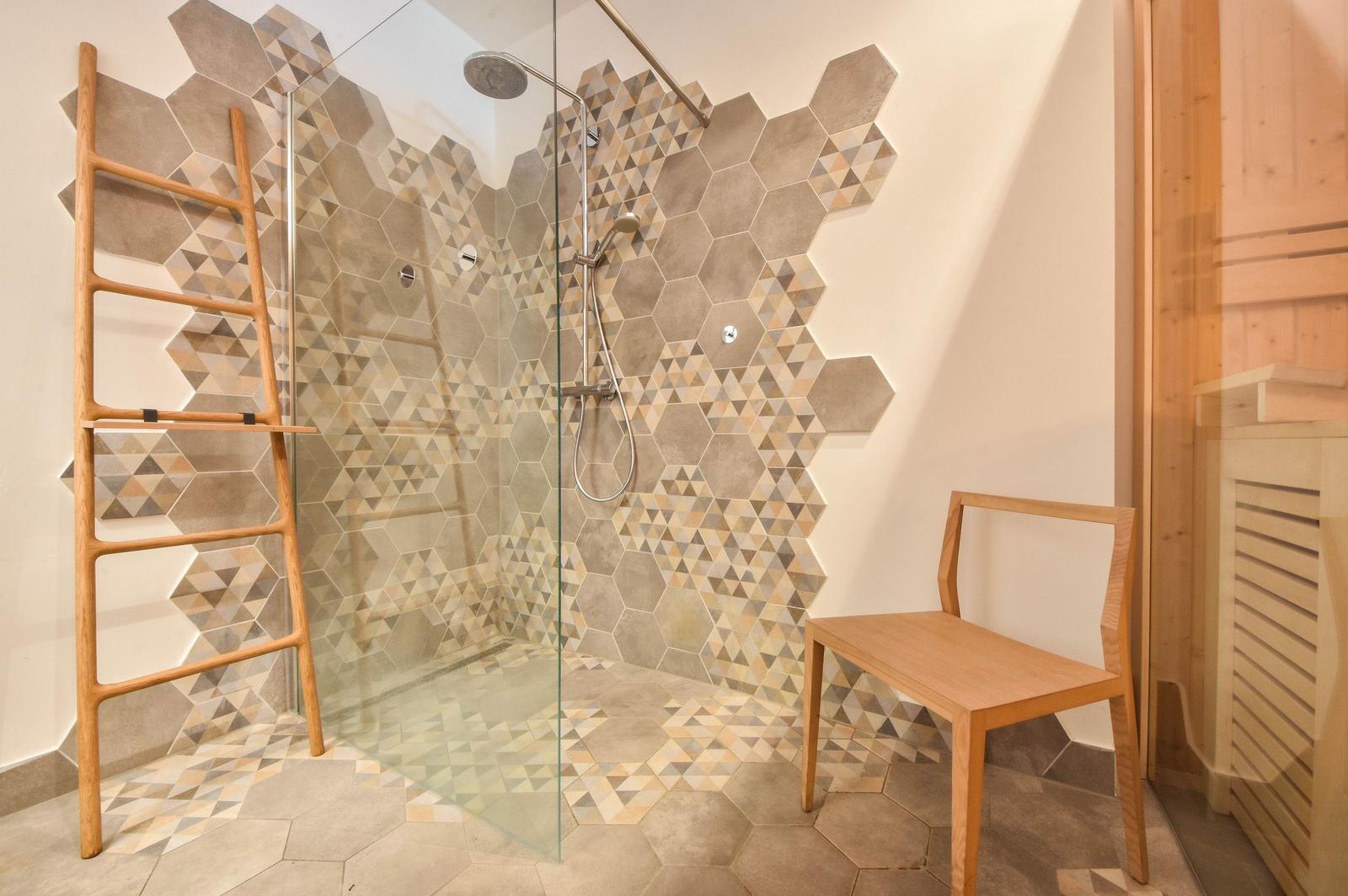 Dusche mit Glaswand, Holzstuhl und Holzleiter, Wand- und Bodenfliesen in Hexagonform.