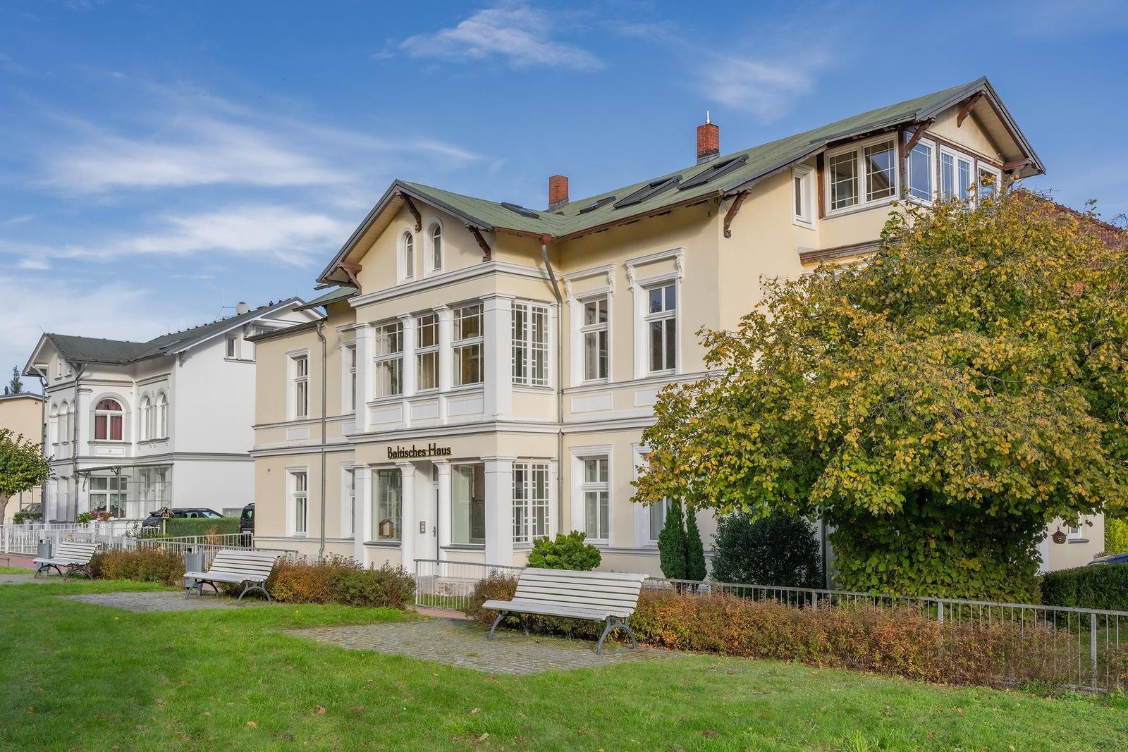 Baltisches Haus mit Garten, Bänken und Bäumen unter blauem Himmel.