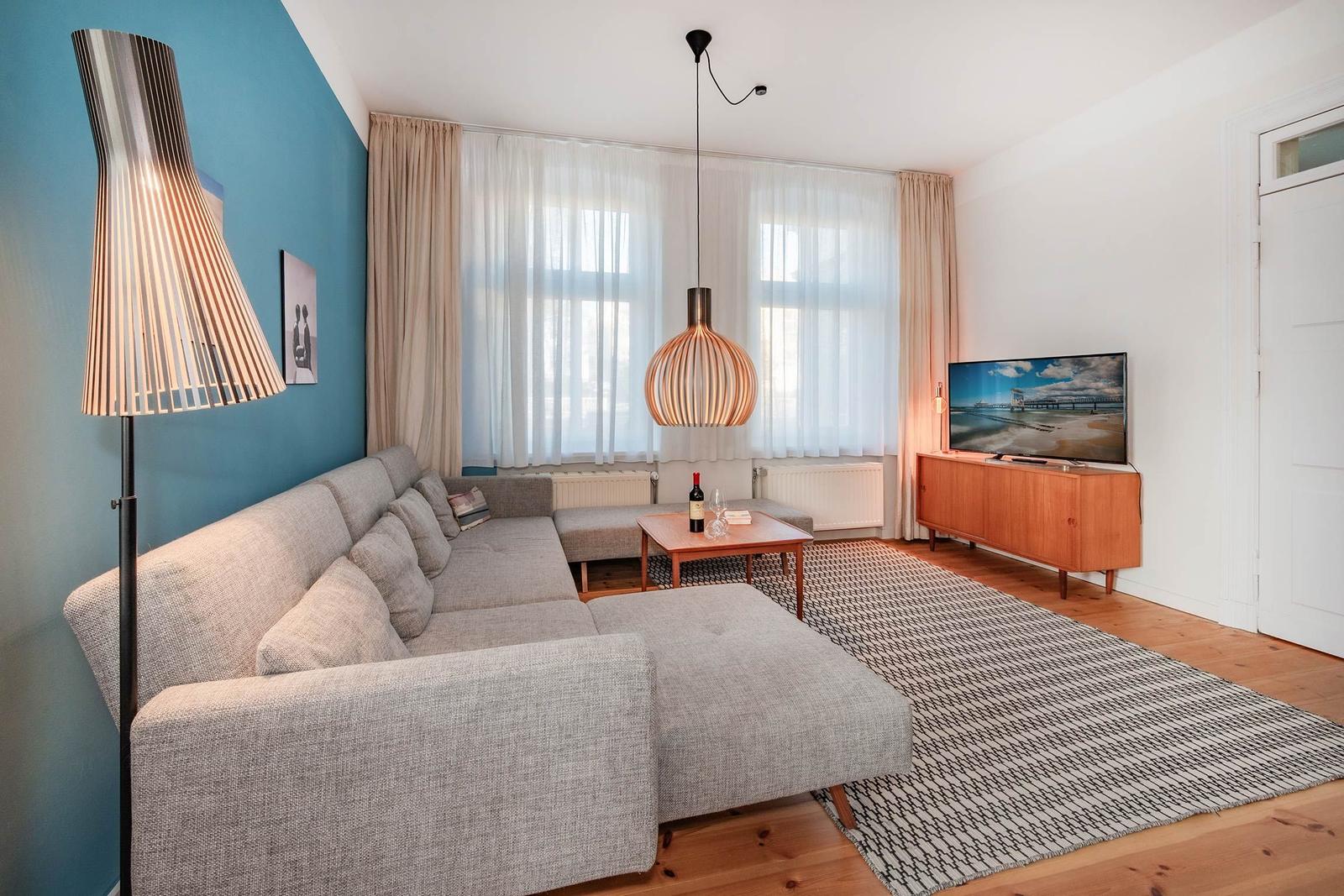 Wohnzimmer mit Sofa, Fernseher und Lampe. Holzboden und Fenster mit Vorhängen.
