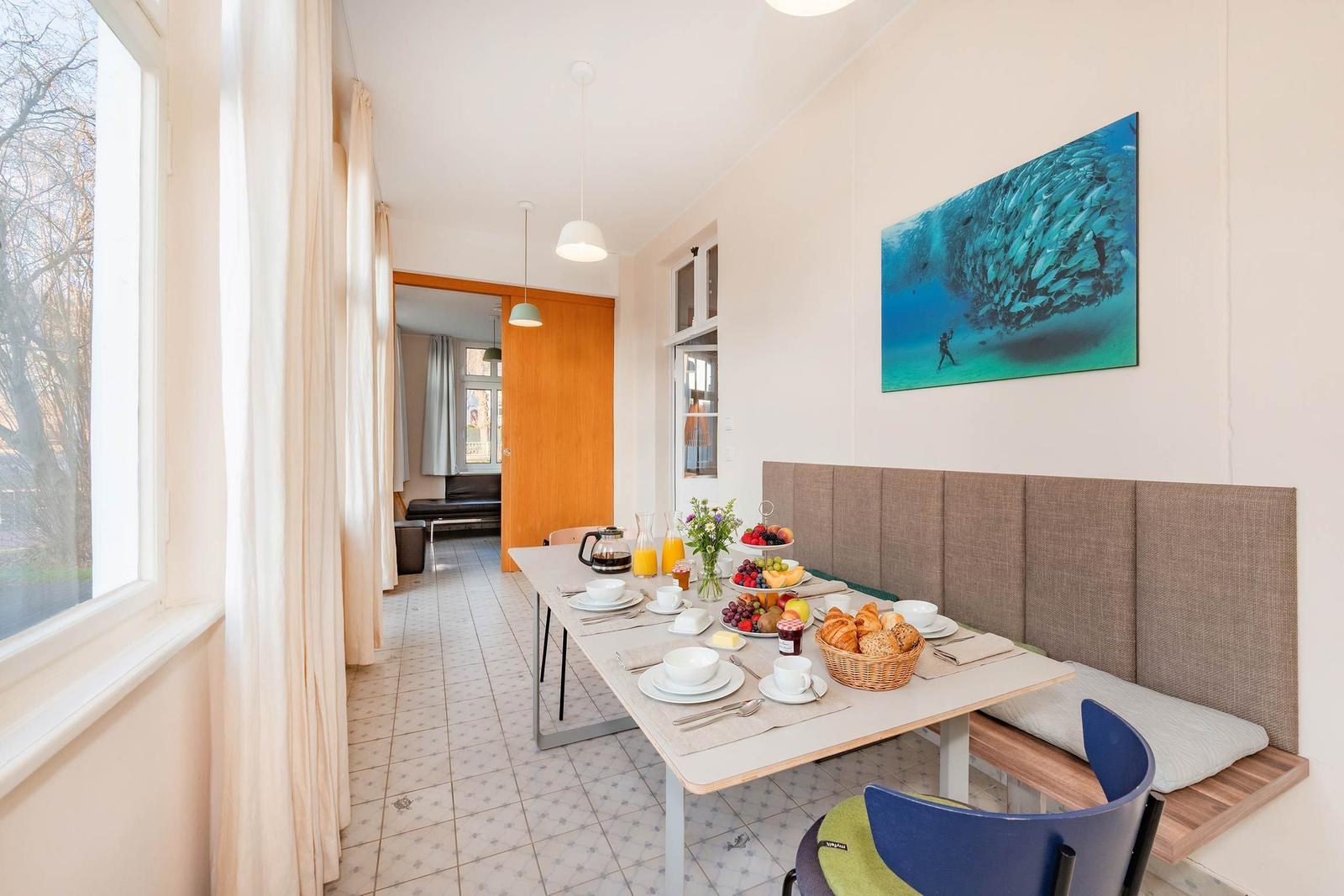 Frühstückstisch mit Obst, Brot und Getränken. Wandbild mit Taucher. Fenster und Sitzbank.
