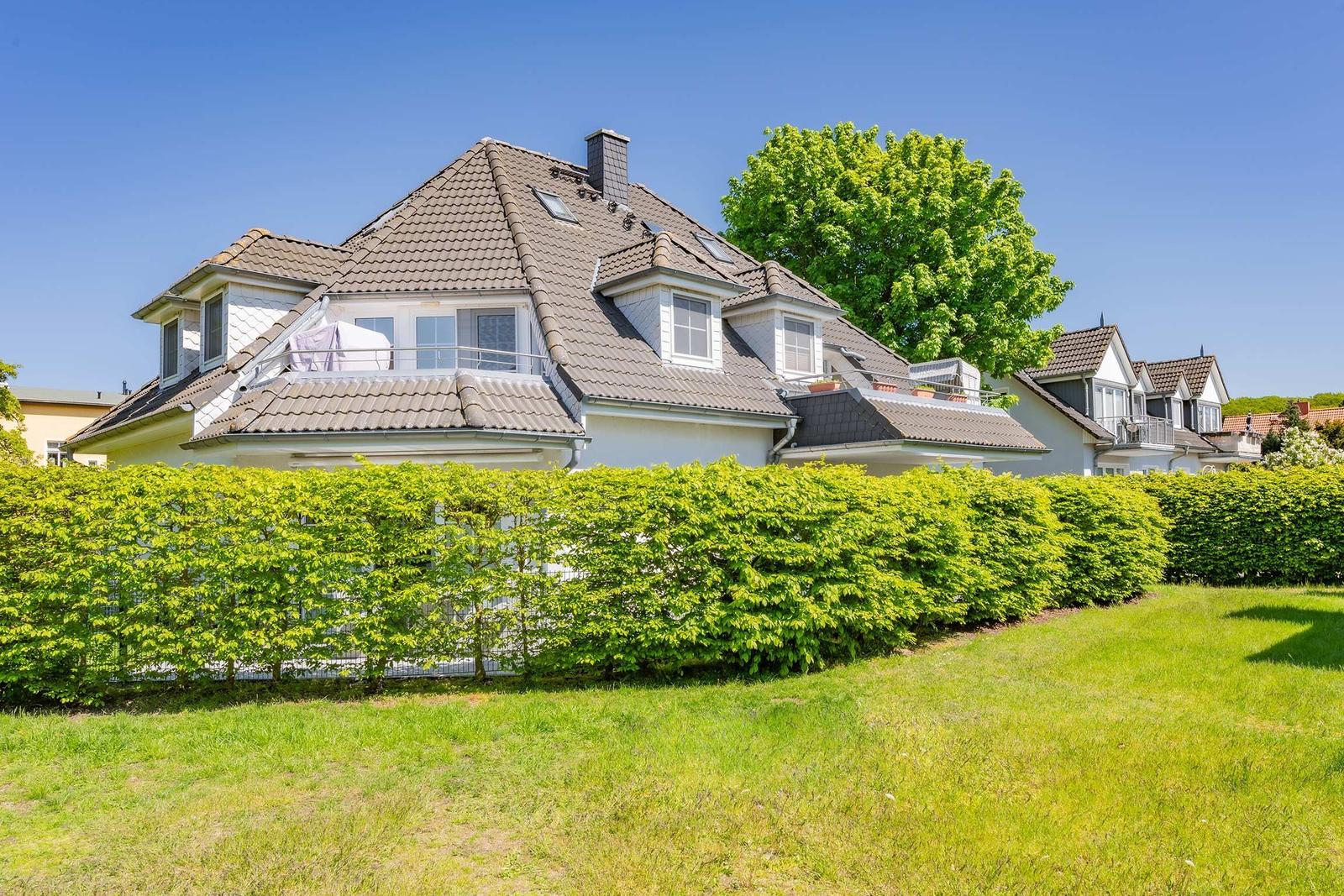 Haus mit Dachgauben und Balkon hinter grünem Heckenzaun.