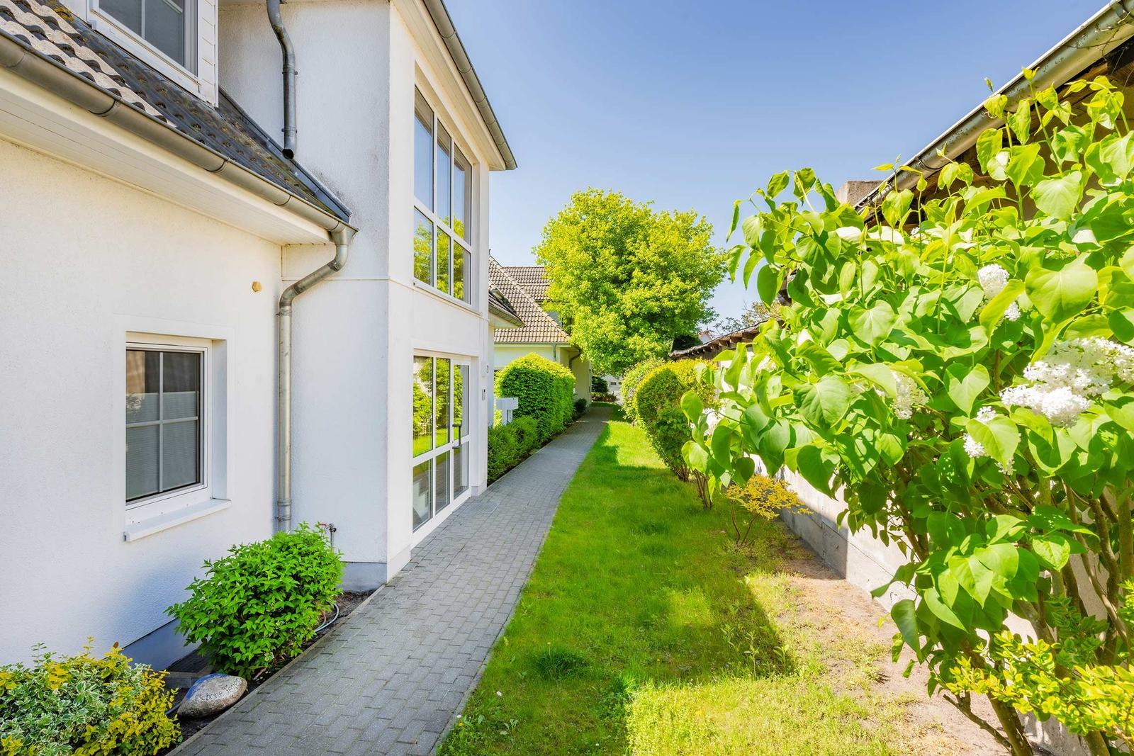 Haus mit gepflasterter Laufstrecke, Rasen und blühenden Sträuchern unter blauem Himmel.