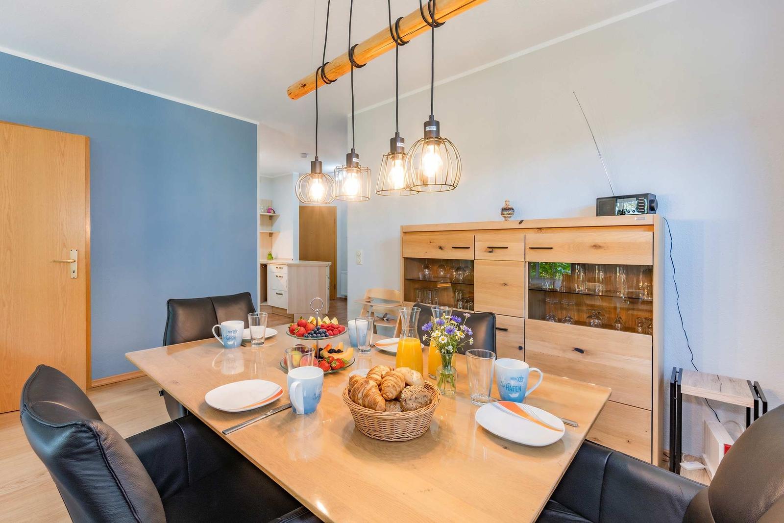 Esszimmer mit Holztisch, schwarzen Stühlen und hängenden Lampen. Auf dem Tisch stehen Frühstückszubehör.