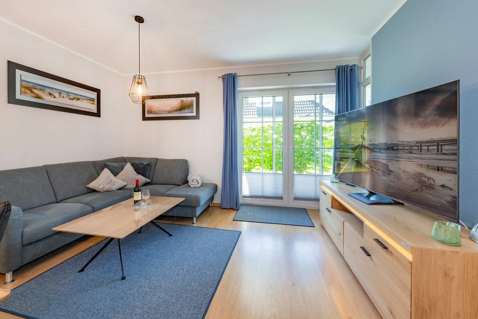 Wohnzimmer mit Sofa, Couchtisch und Fernseher. Blick auf grüne Bäume durch Fenster.