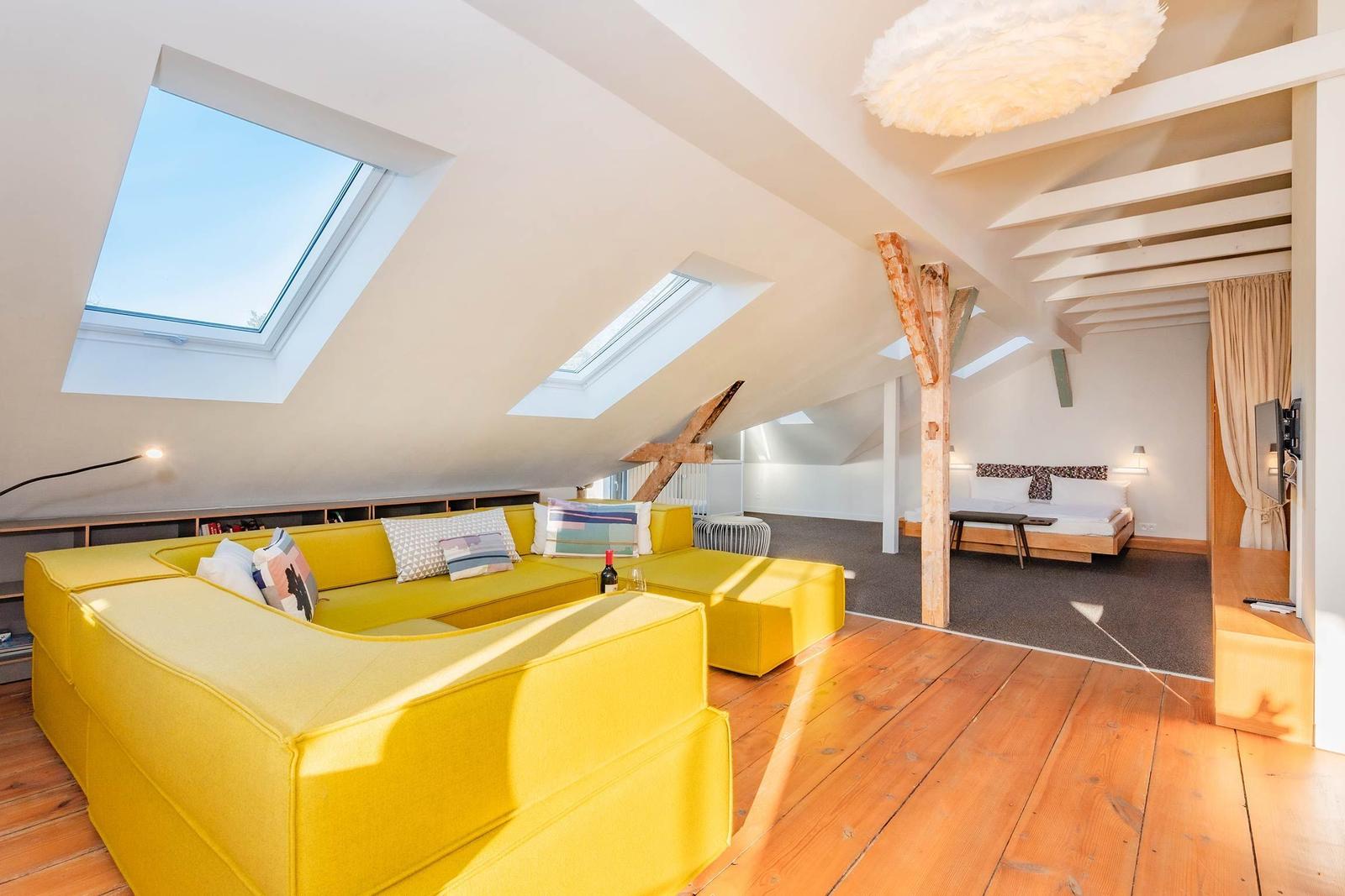 Ein Schlafzimmer mit gelbem Sofa, Holzboden und Dachfenstern. Im Hintergrund befindet sich ein Bett.