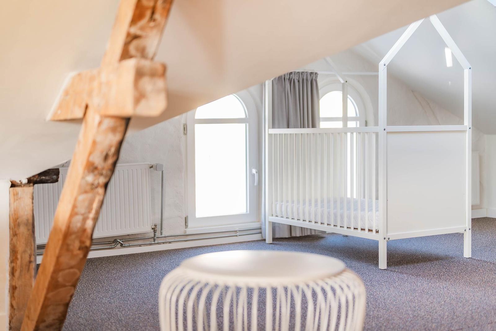 Ein Kinderzimmer mit weißem Babybett und Holzelementen. Fenster mit grauen Vorhängen.