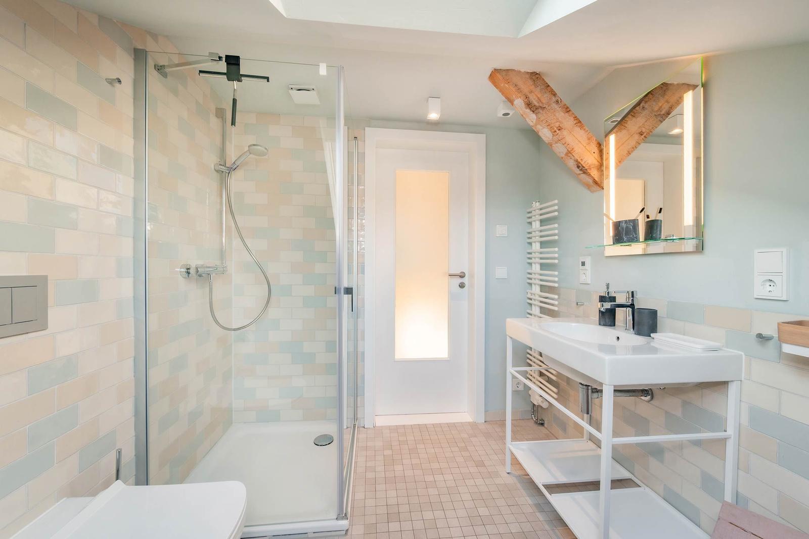 Badezimmer mit Dusche, Waschbecken und Toilette. Holzdeckenbalken und gefliesten Wänden.