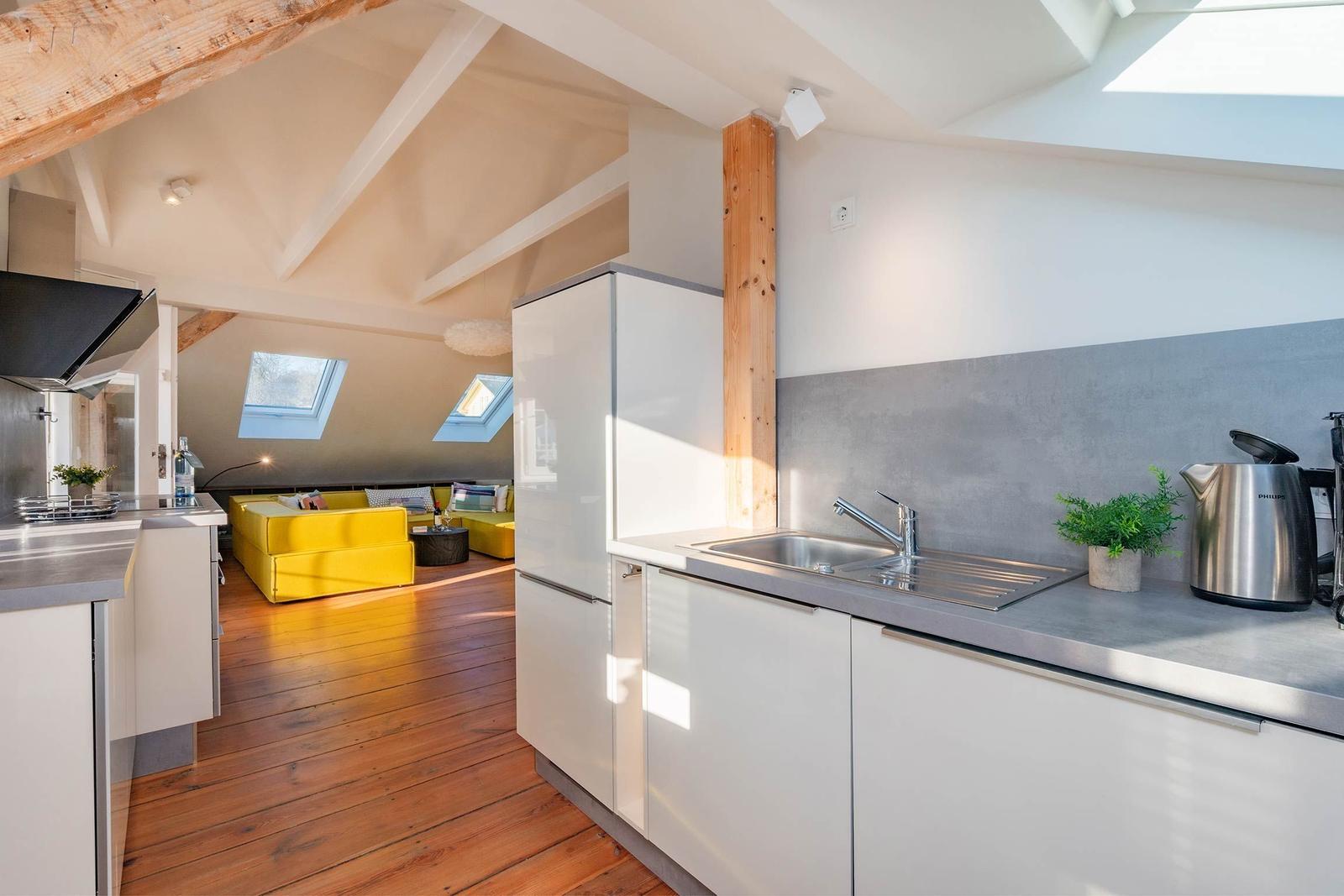 Küche mit Arbeitsfläche, Spüle und Kühlschrank. Im Hintergrund Wohnbereich mit gelbem Sofa.