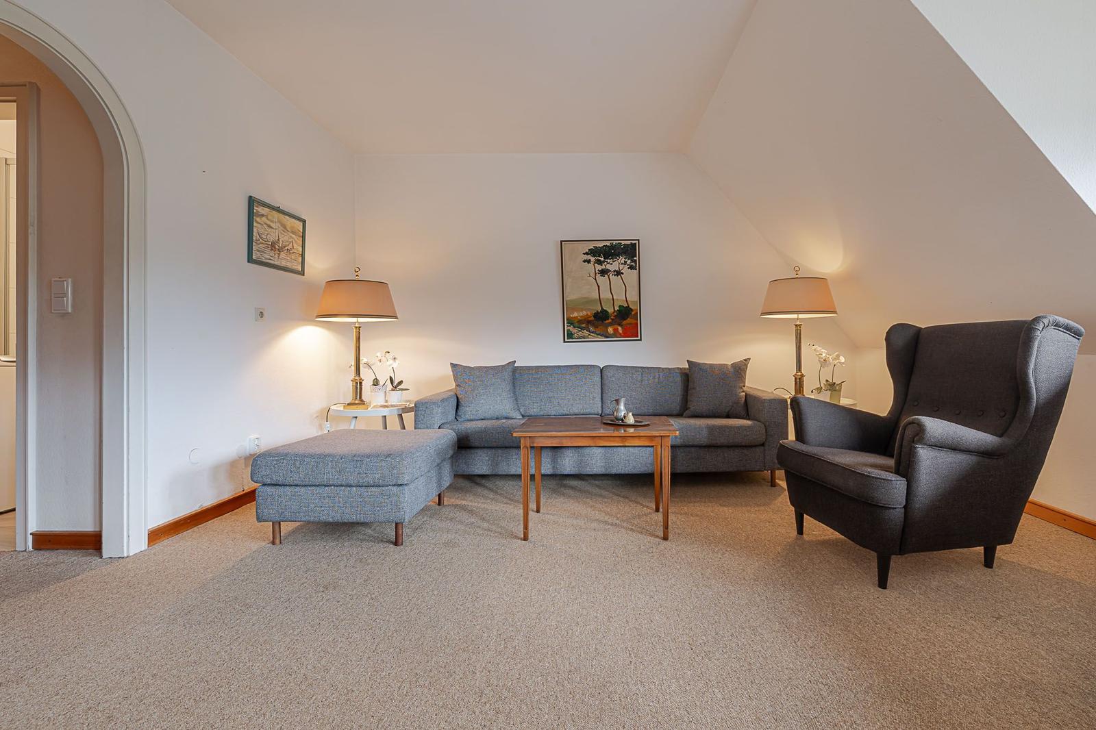 Wohnzimmer mit Sofa, Sessel und Tisch unter schrägen Dachdecken.