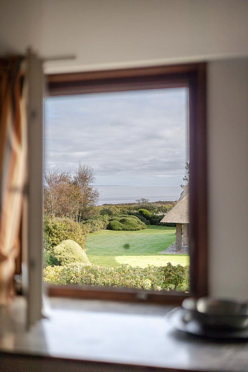 Durch das Fenster blickt man auf einen grünen Garten und das Meer.