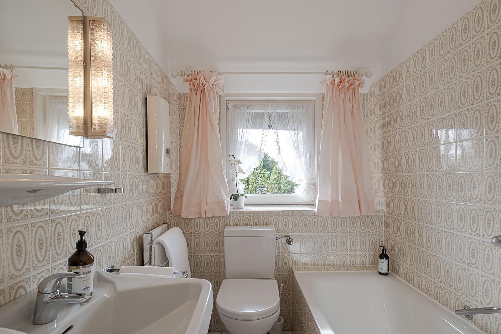 Badezimmer mit Badewanne, Toilette und Waschbecken. Wandfliesen mit Muster und Fenster mit rosa Vorhängen.