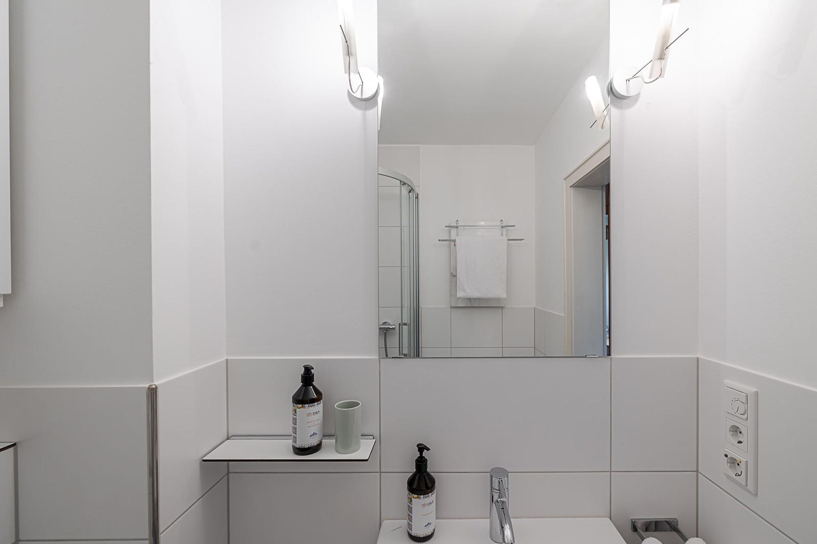 Badezimmer mit Spiegel, Dusche und Waschbecken. Weiße Wände und moderne Beleuchtung.