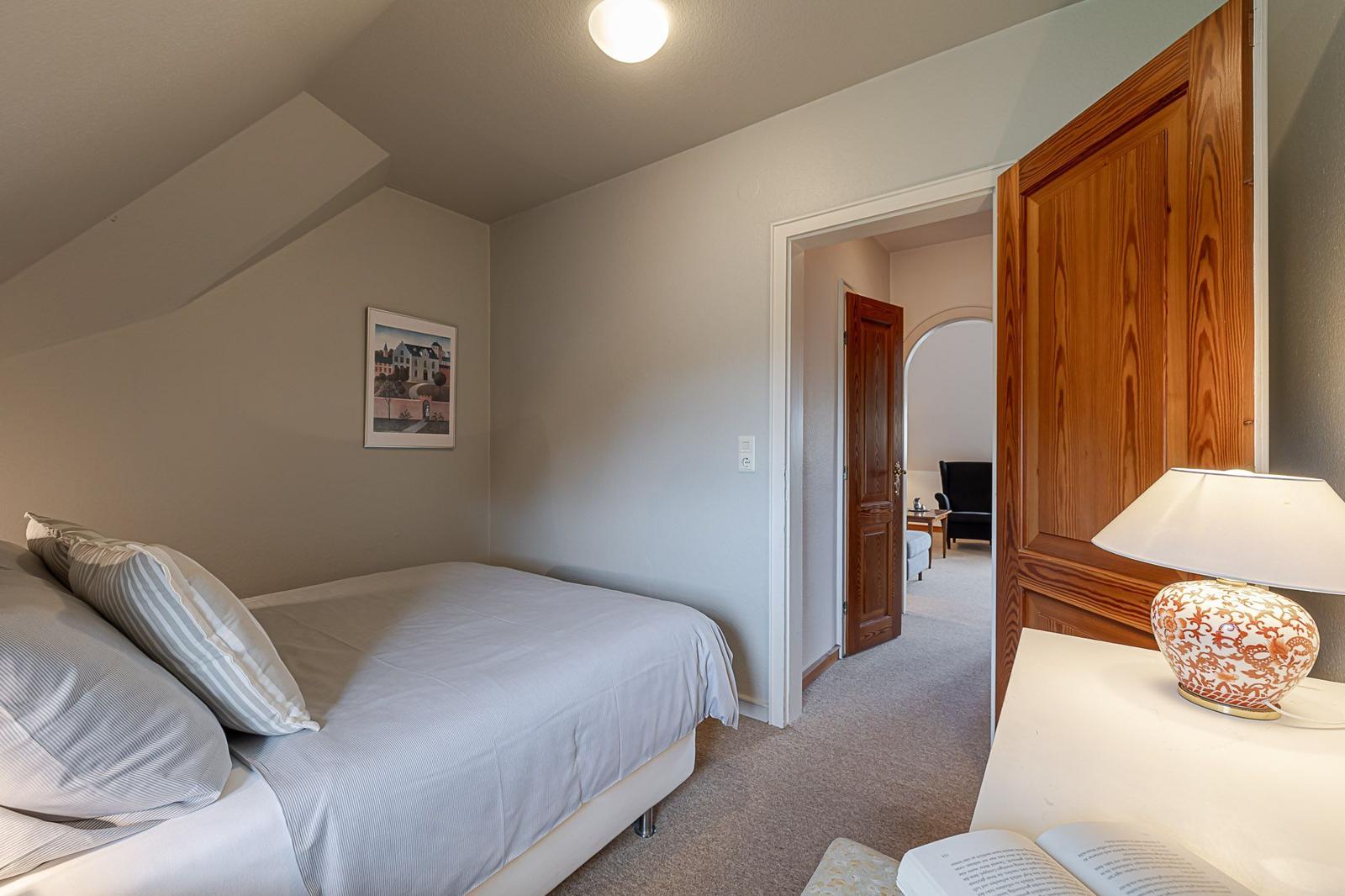 Schlafzimmer mit Bett, Nachttisch und Lampe. Durchgang zu Wohnbereich mit Sitzgelegenheit.