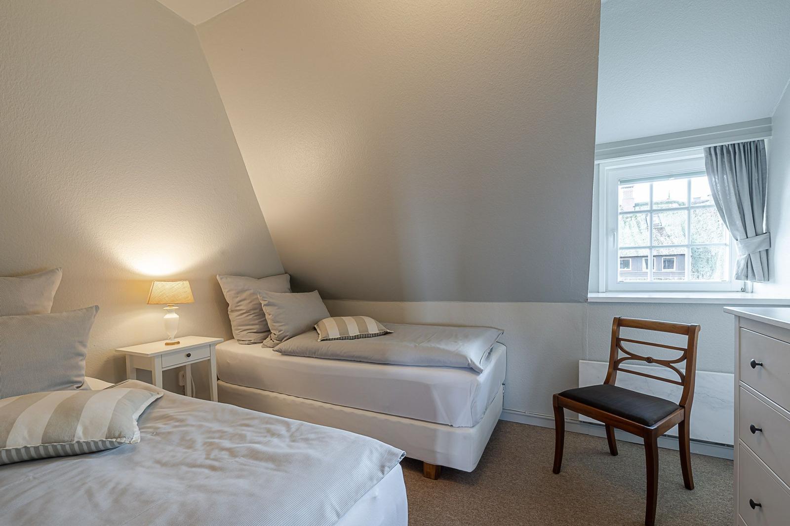 Zimmer mit zwei Betten, Nachttisch, Stuhl und Fenster mit Vorhang.