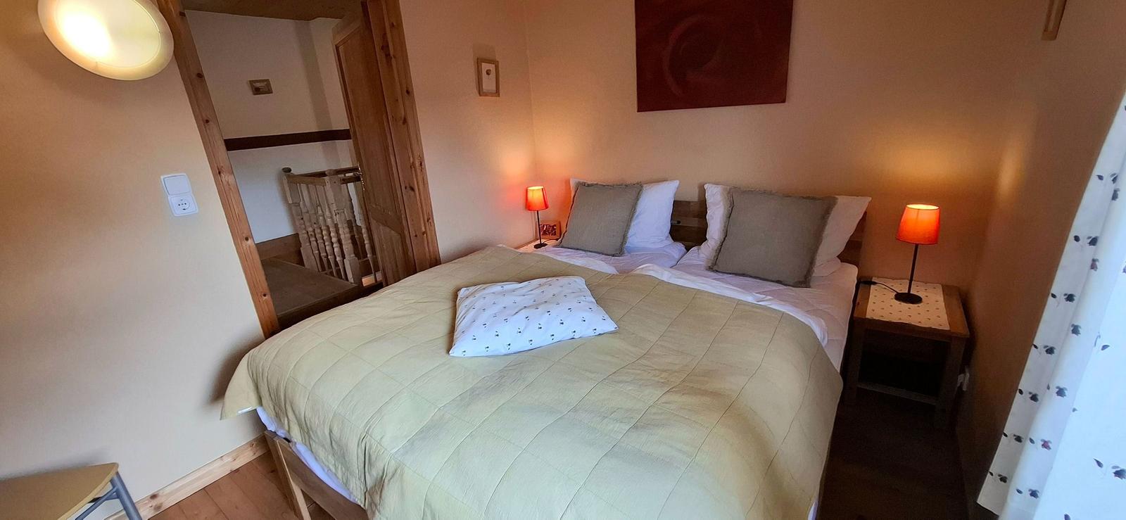 Schlafzimmer mit Doppelbett, zwei Nachttischen und Lampe. Hinter dem Bett ist eine Tür sichtbar.
