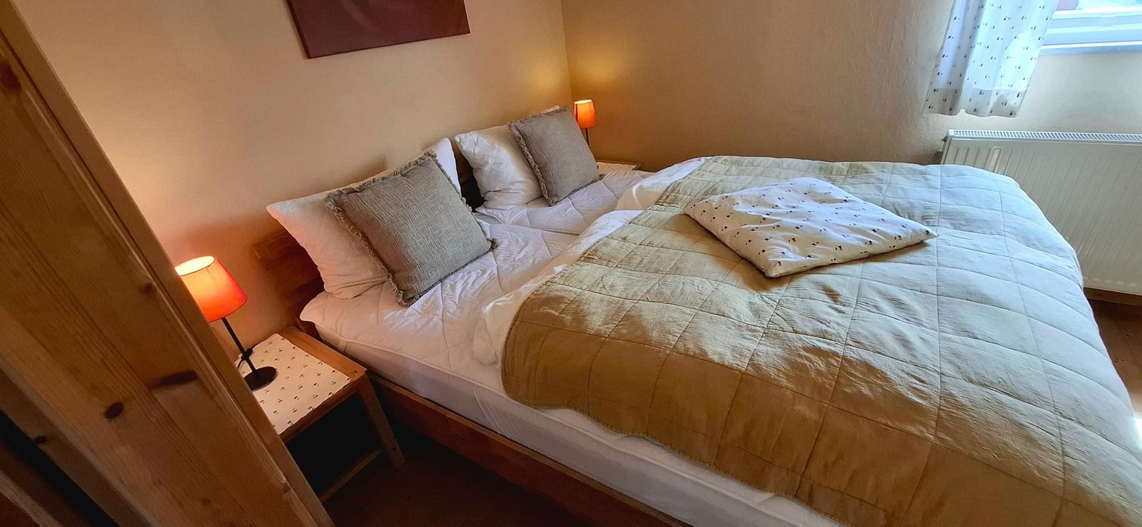 Doppelbett mit Decke und Kissen, zwei Nachttische mit Lampen und Fenster mit Vorhang.