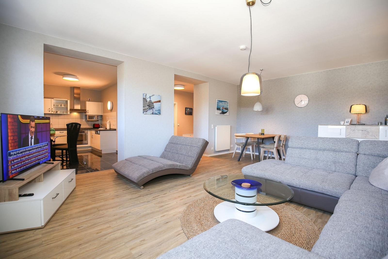 Wohnzimmer mit Sofa, Couchtisch und Fernseher. Küche und Essbereich sind sichtbar.