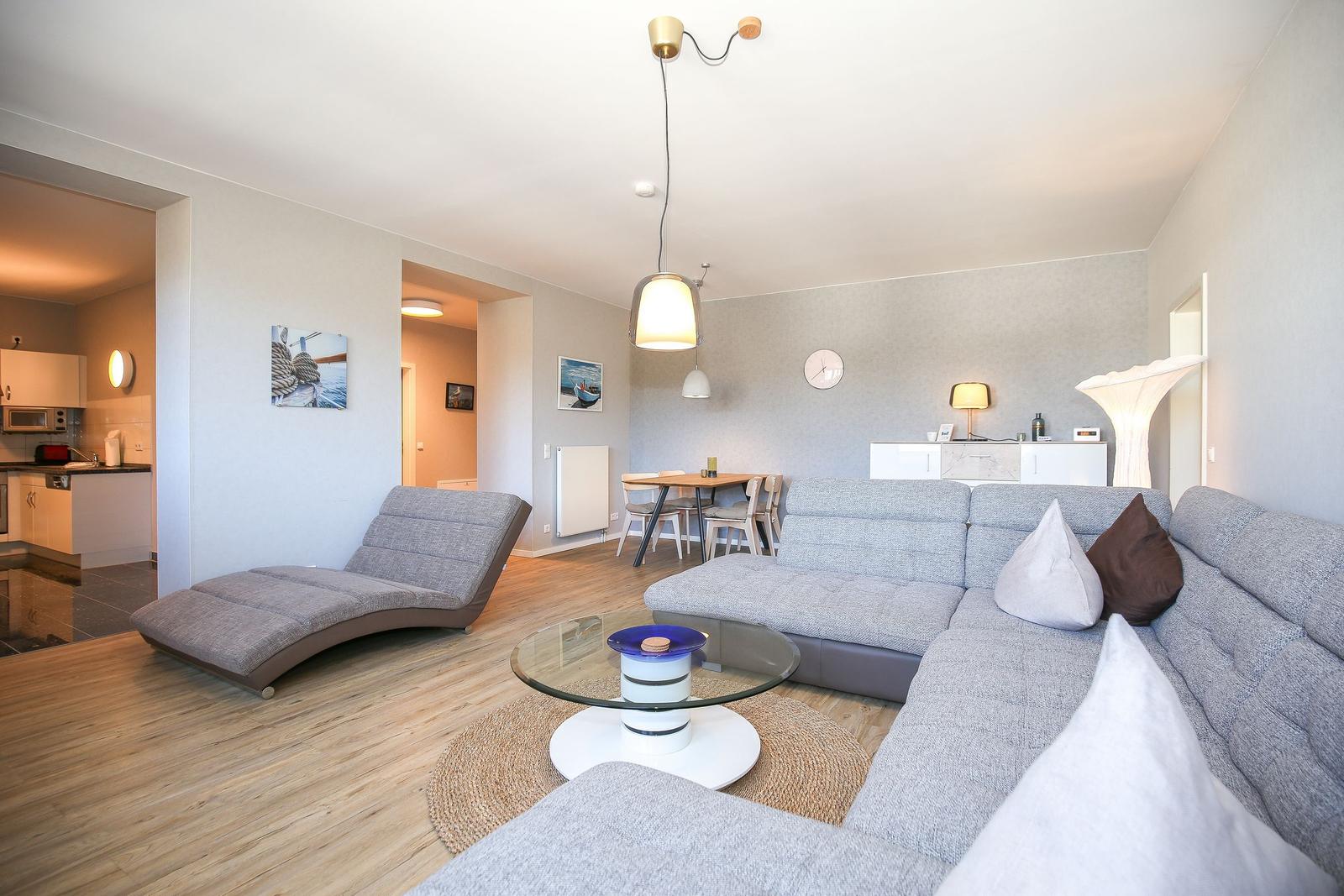 Wohnzimmer mit grauem Sofa, Glas-Tisch und Holzboden. Küche und Essbereich sind sichtbar.