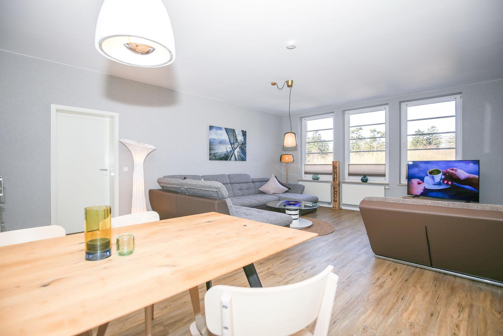 Wohn- und Essbereich mit Sofa, Tisch und TV. Holzboden und große Fenster.