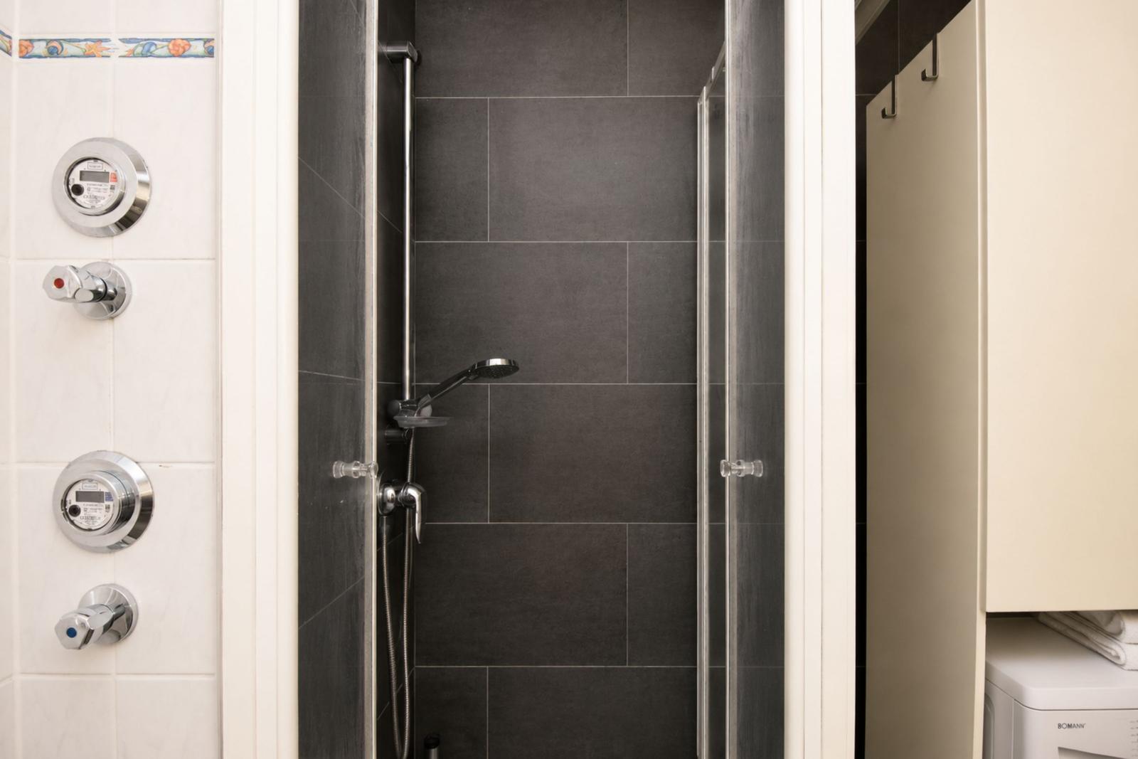 Dusche mit Glaswand, dunklen Fliesen und Chromarmaturen. Links Thermostat und Wasserhähne.