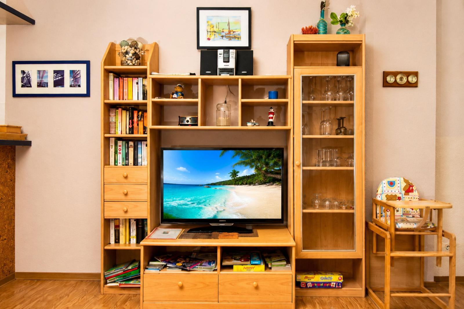 Holzregal mit Fernseher, Büchern und Gläsern. Kinderhochstuhl und Wandbilder sind sichtbar.