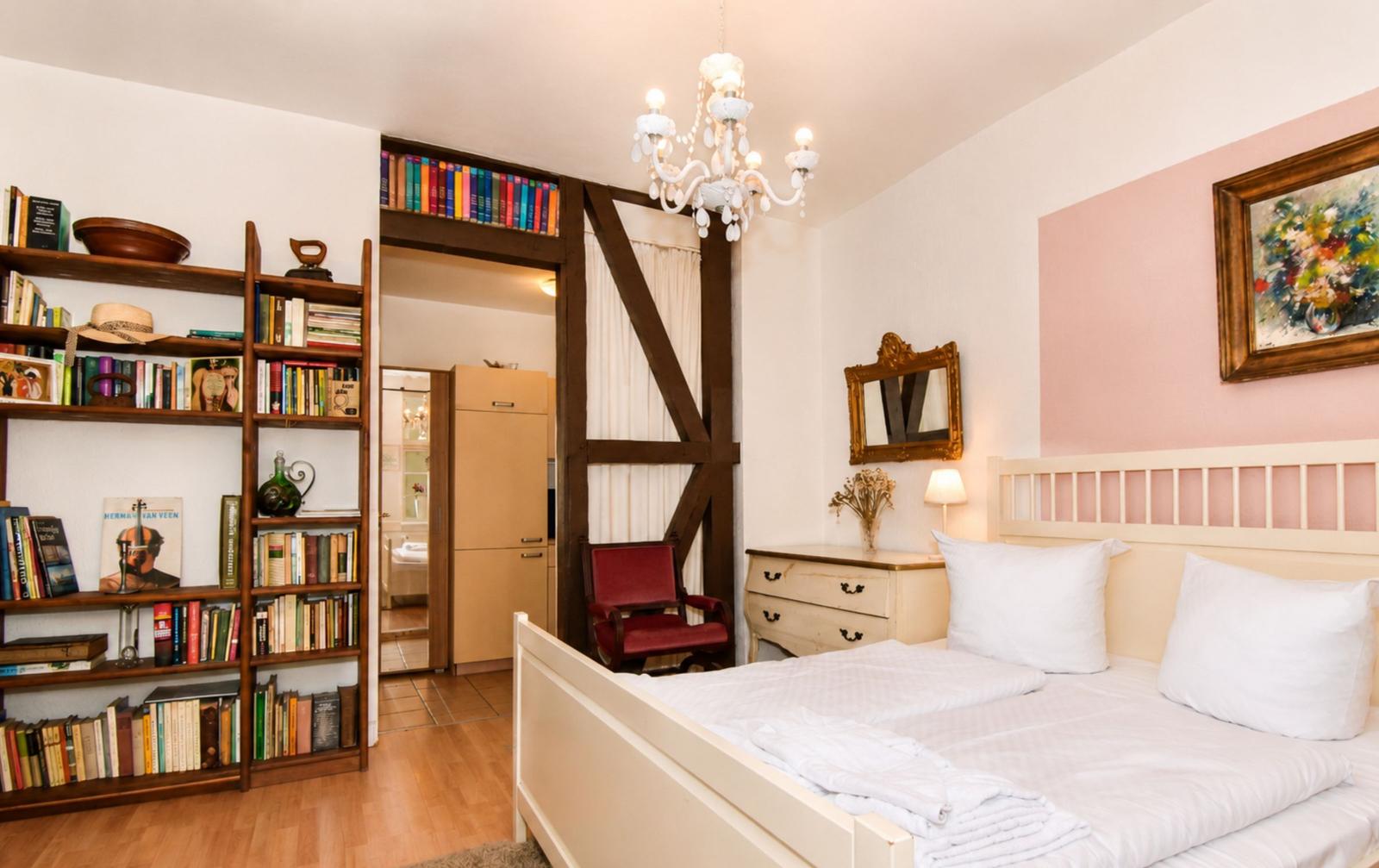 Schlafzimmer mit Bett, Bücherregal, Spiegel und Kronleuchter. Durchgang zu Bad und Wohnbereich.