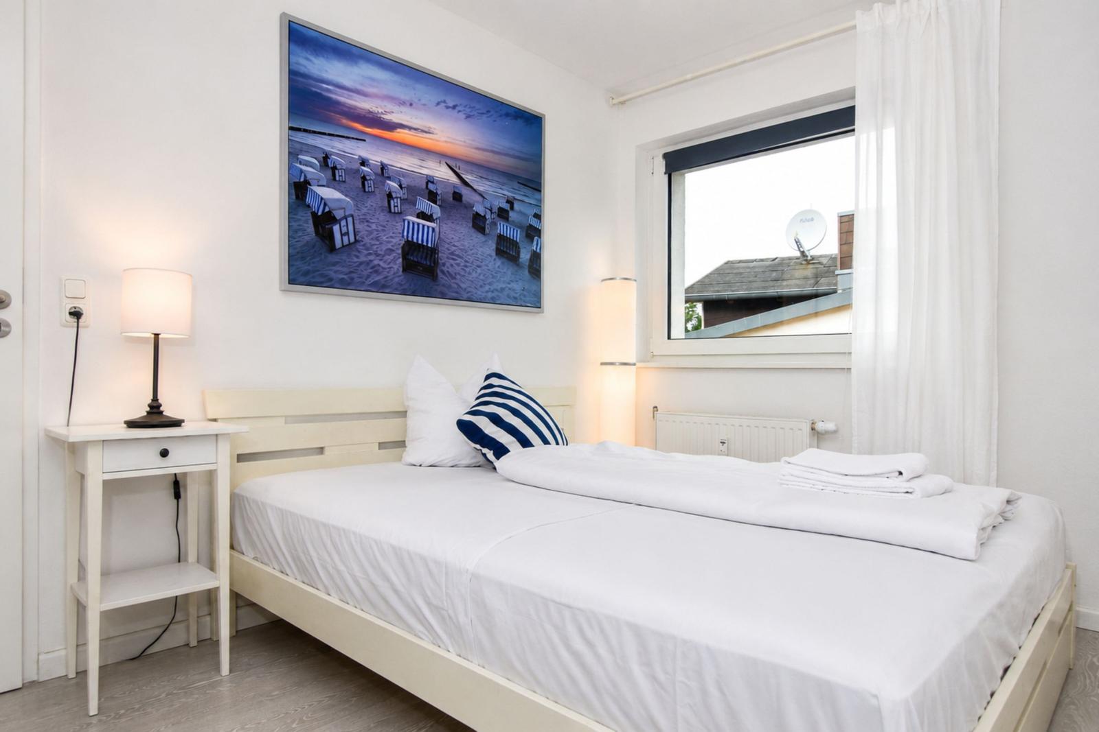 Schlafzimmer mit Bett, Nachttisch und Wandbild. Fenster mit weißen Vorhängen.