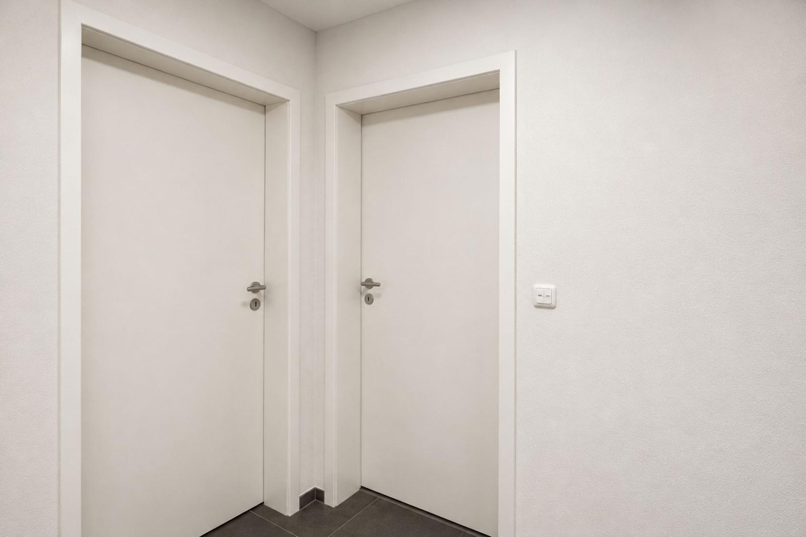 Zwei weiße Türen mit silbernen Griffen und einem Lichtschalter an der Wand.