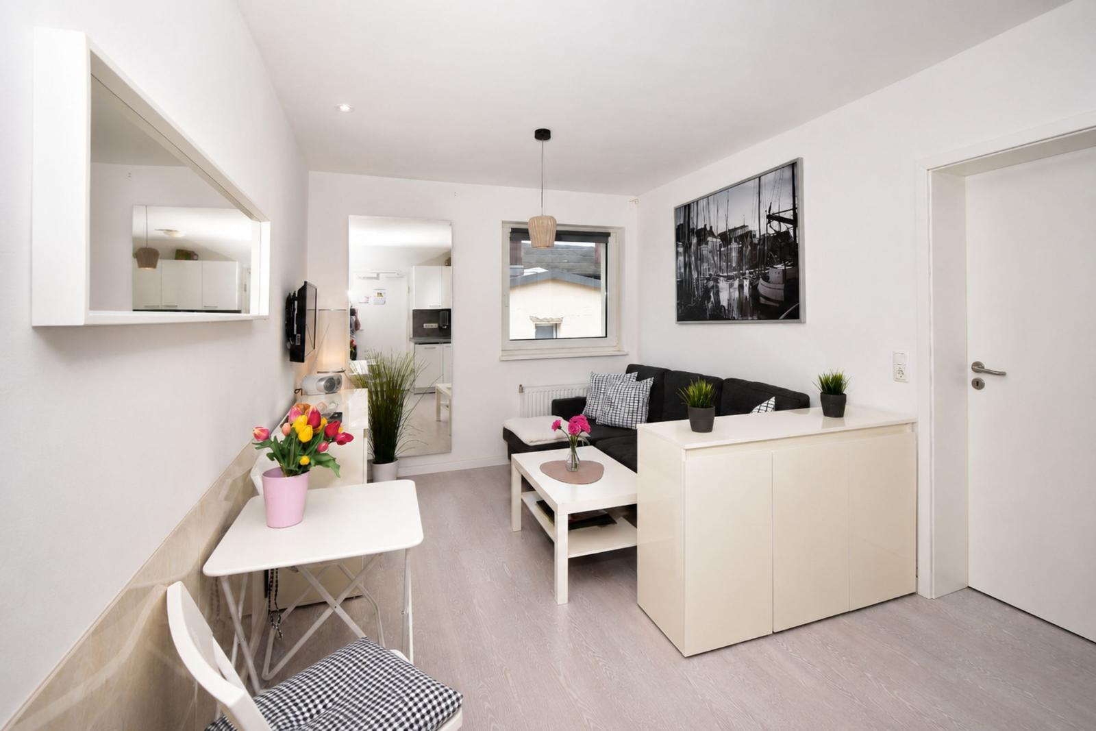Wohnbereich mit Sofa, Tisch und Wandbild. Küche im Hintergrund sichtbar.