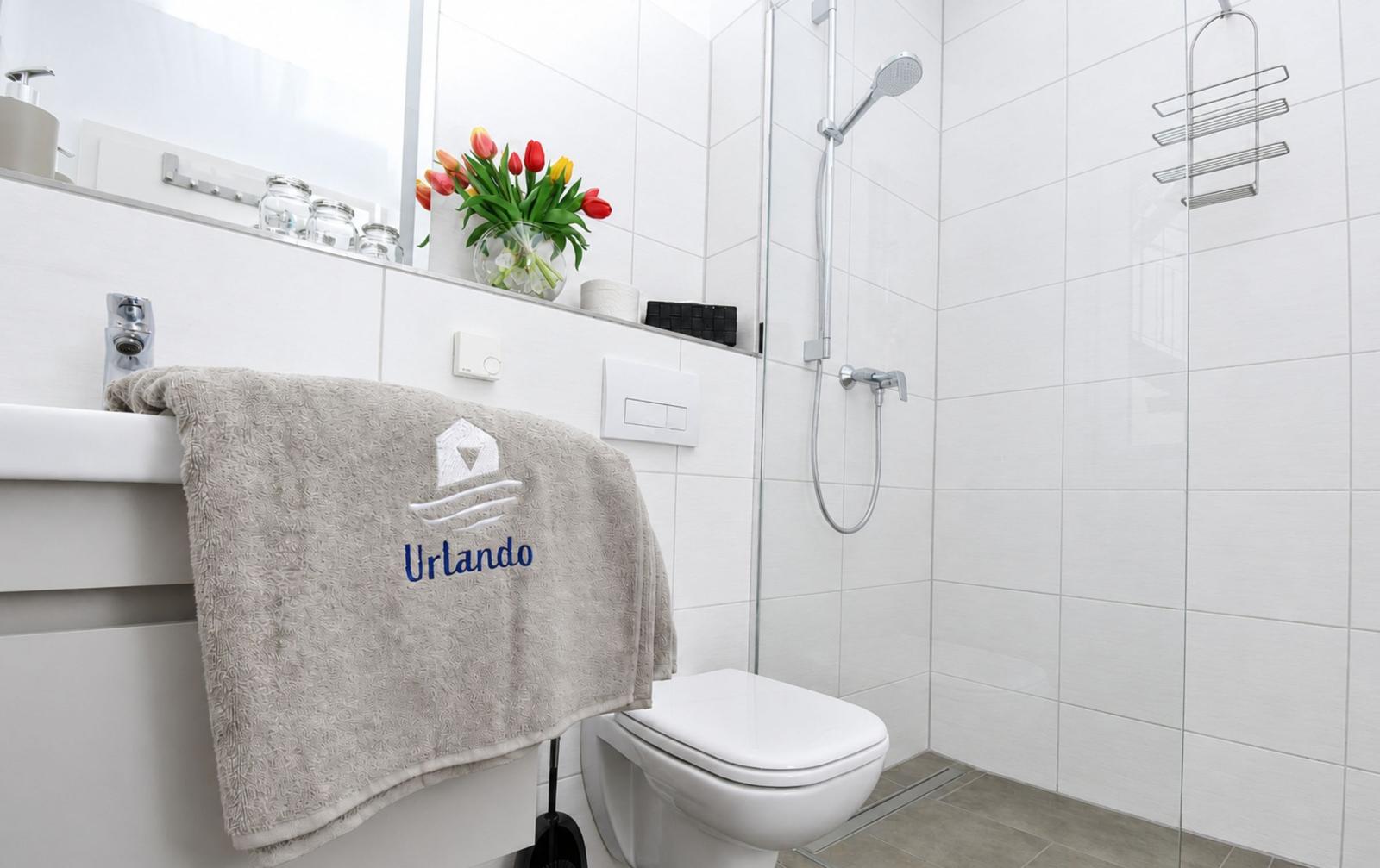 Badezimmer mit Dusche, Toilette und Waschbecken. Handtuch mit Logo liegt über Waschbecken.