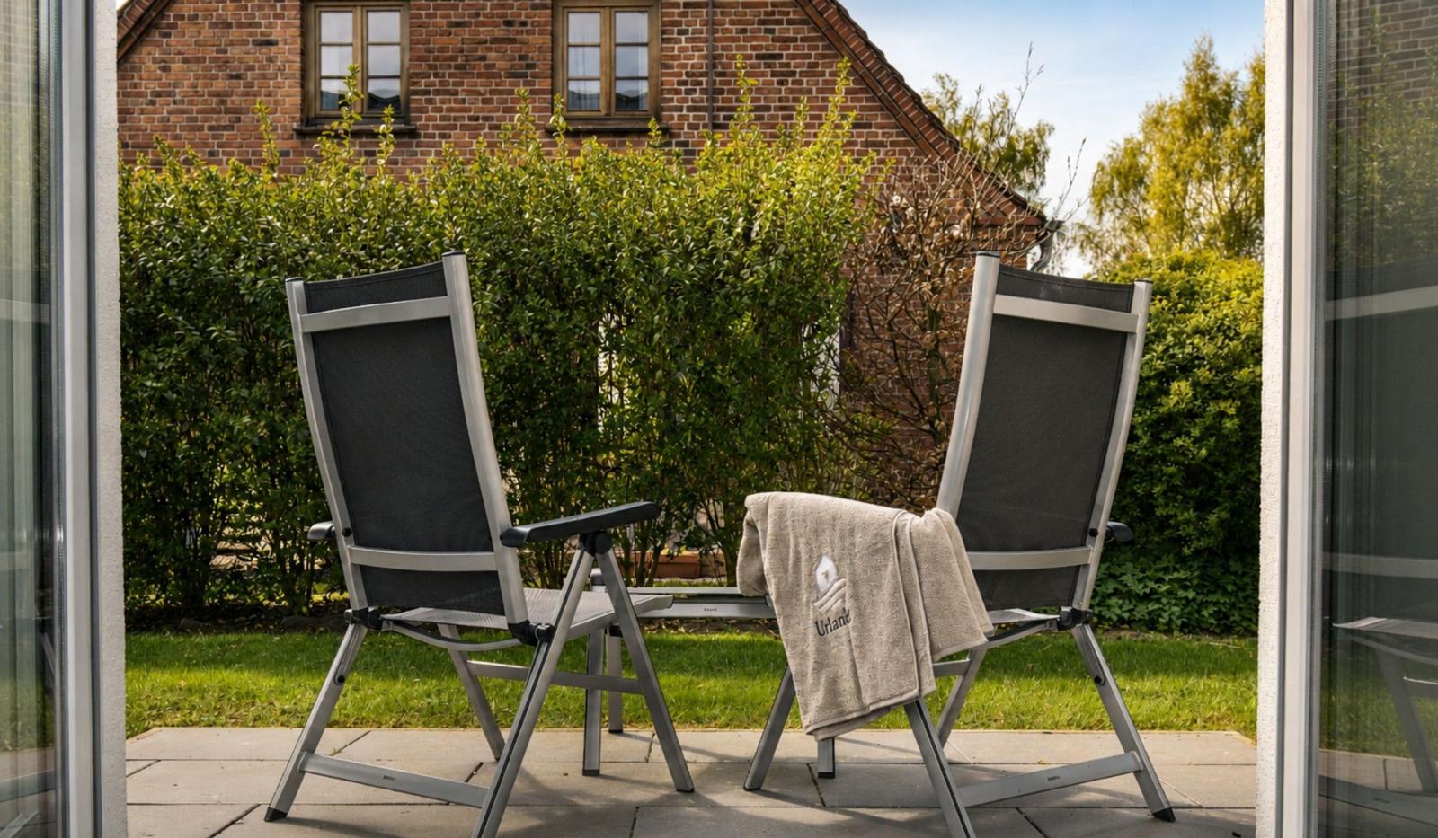 Zwei Stühle auf einer Terrasse mit Blick auf einen Garten und ein Backsteinhaus.