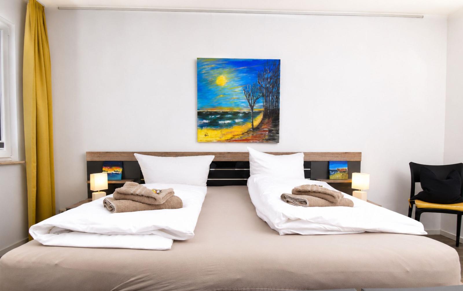 Zwei Betten mit weißen Laken und Handtüchern. Wandbild und gelbe Vorhänge.