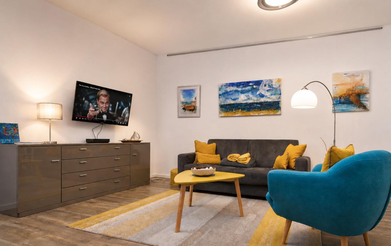 Wohnzimmer mit Sofa, blauer Sessel und Fernseher an Wand.