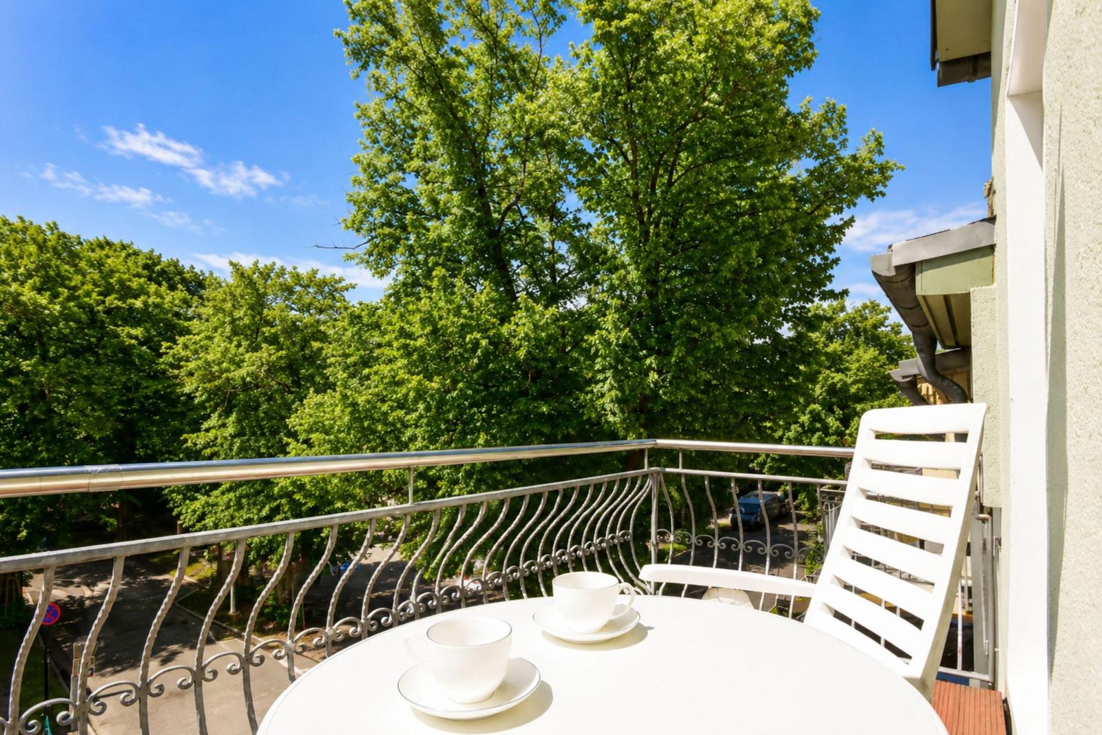 Balcony with white table and chairs, metal railing, and view of green trees under blue sky.
