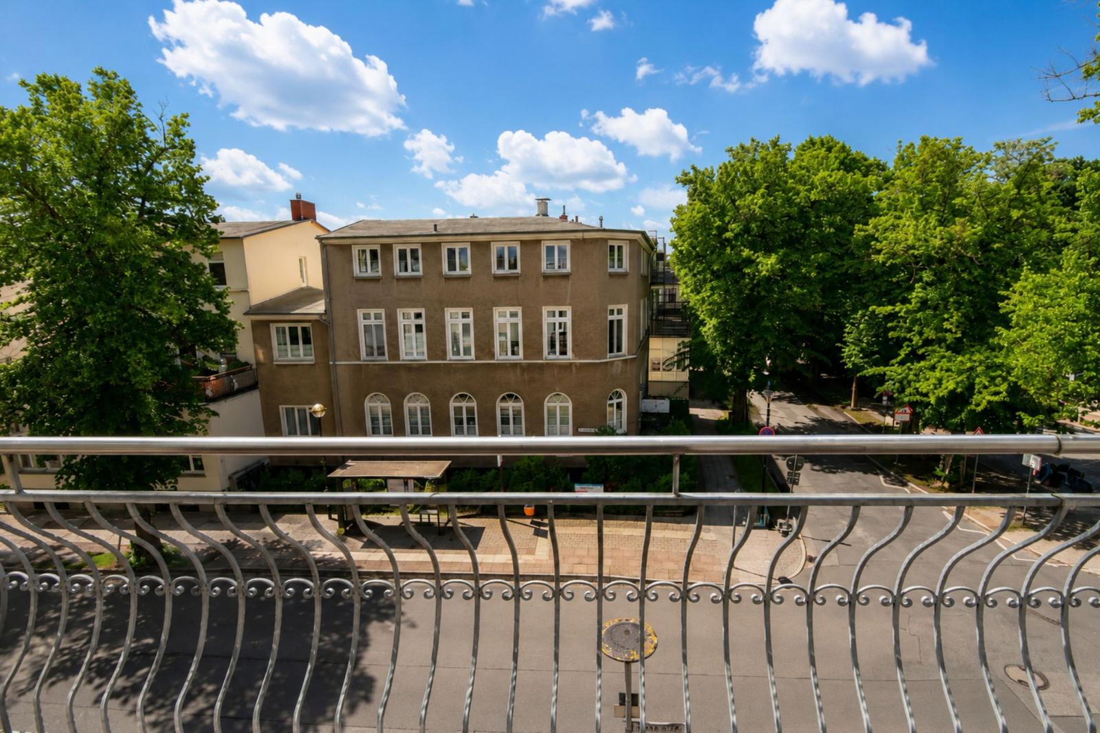 Blick vom Balkon auf ein Haus mit weißen Fenstern und Bäumen unter blauem Himmel.