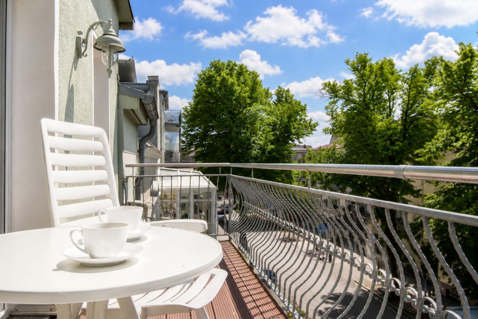 Balcony with white table and chairs, metal railing, and view of trees and buildings.