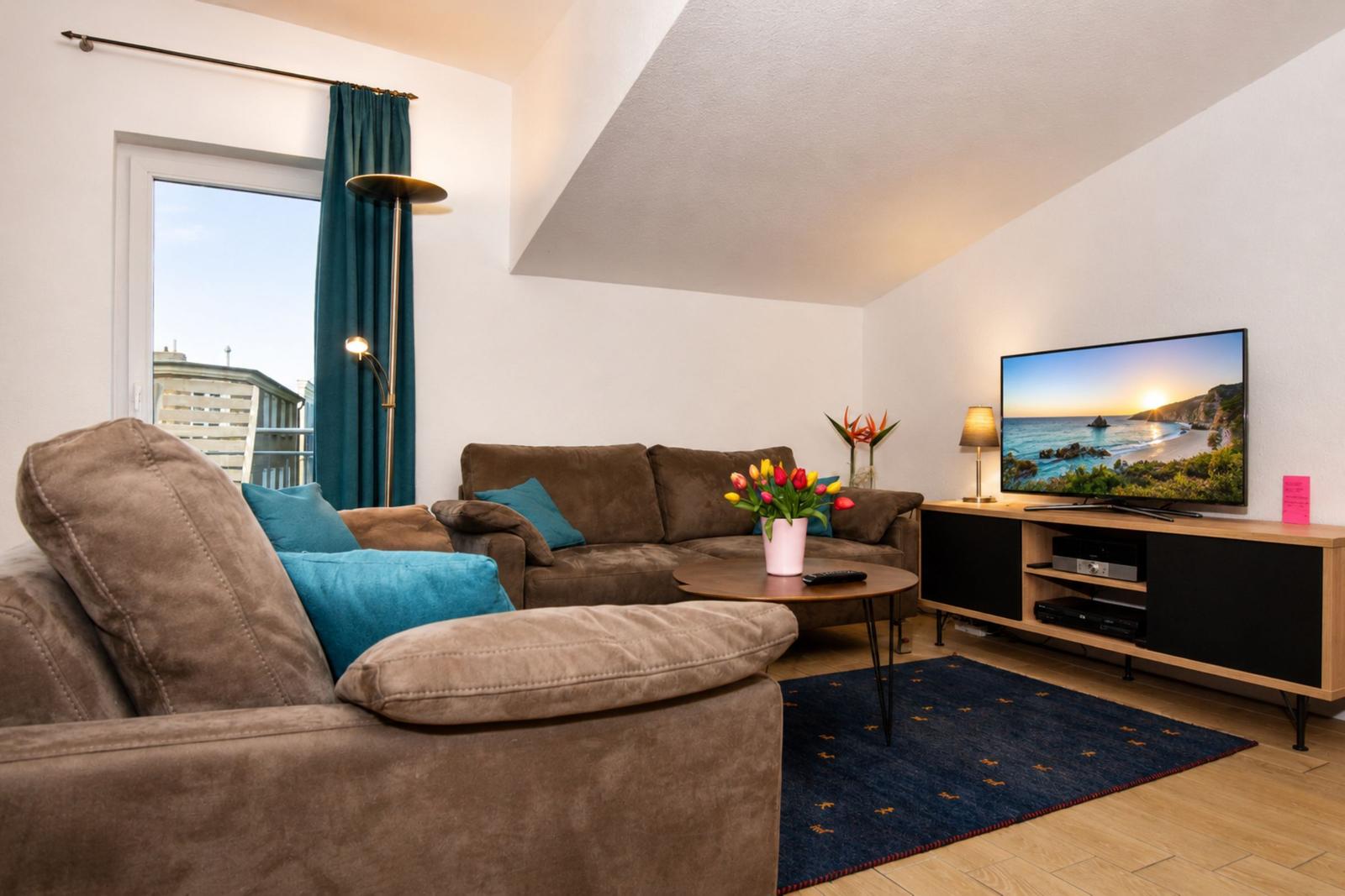 Ein Wohnzimmer mit braunem Sofa, blauem Teppich und Fernseher auf Holzregal.
