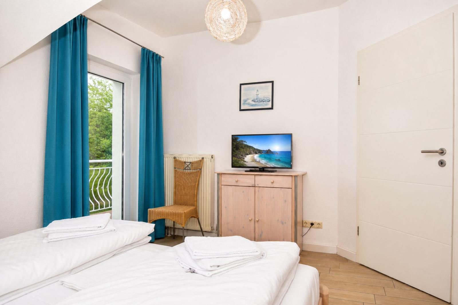 Schlafzimmer mit Bett, TV und Balkon. Blaue Vorhänge und Holzmöbel.